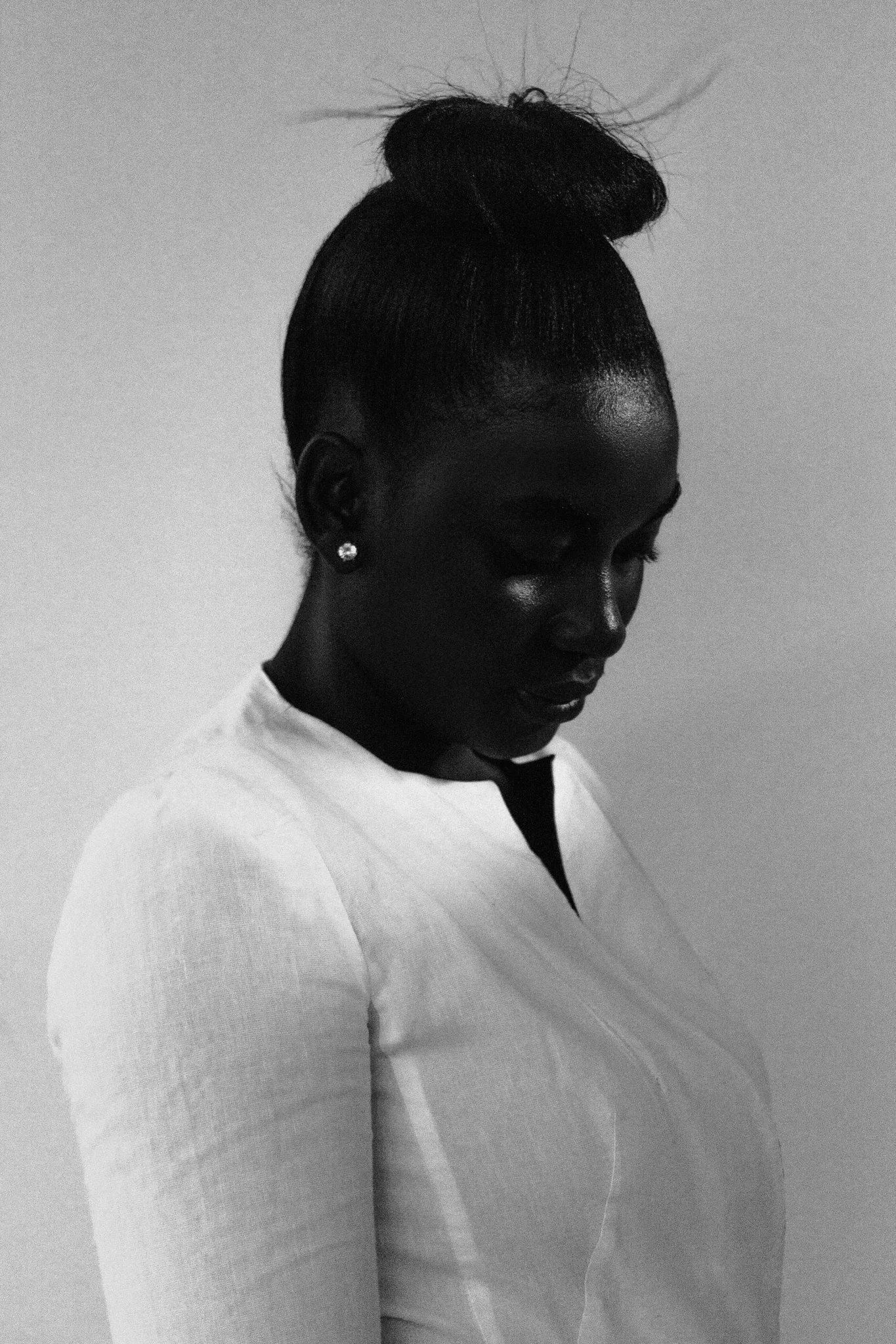 A high-contrast, black and white fine art portrait of a woman with her hair styled in a high bun, wearing earrings and a light-colored top against a plain background.