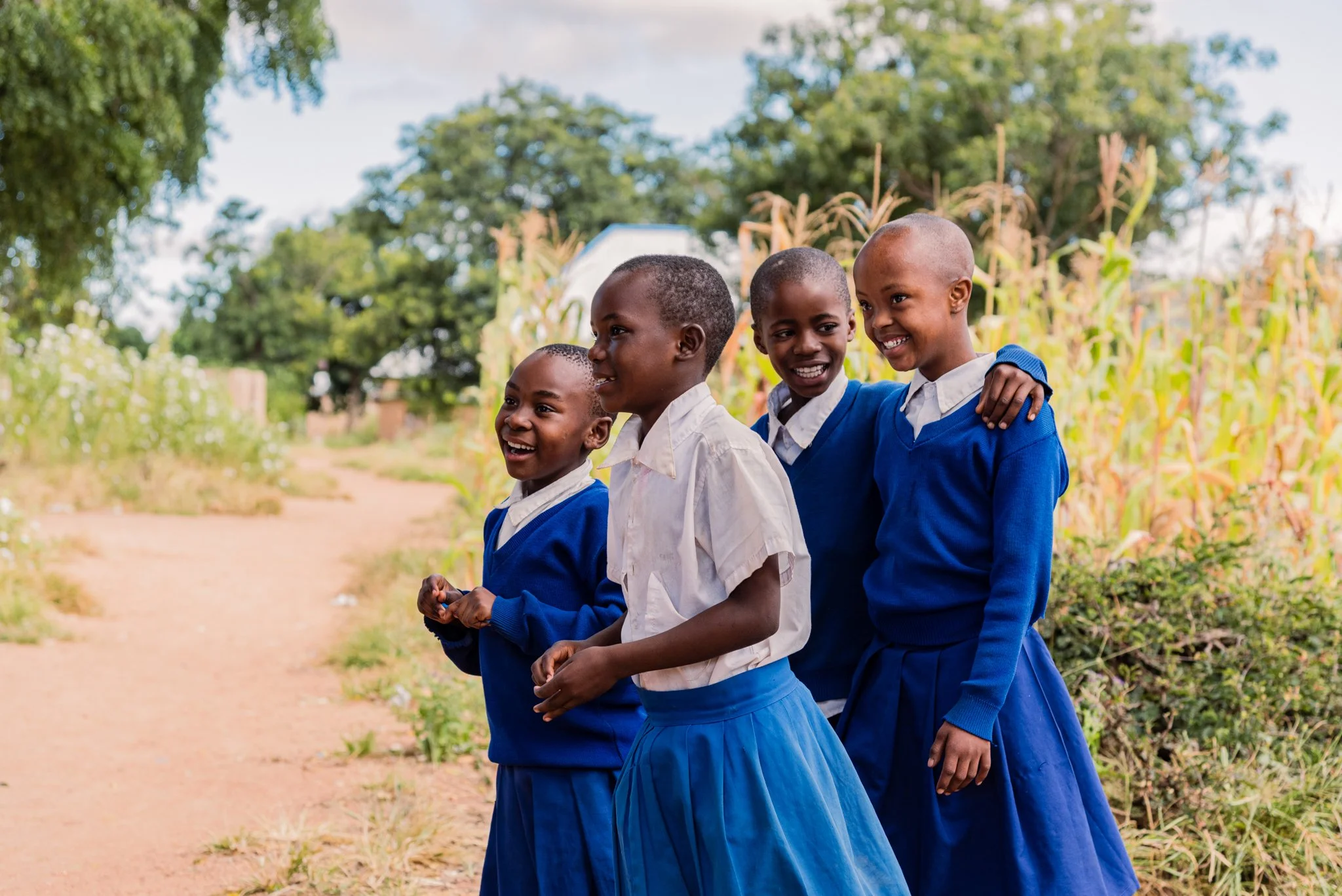 Four children in blue school uniforms walking outdoors on a dirt path at a primary school in Dodoma, Tanzania, with trees and vegetation in the background, during an inclusive education programme supported by Leonard Cheshire.