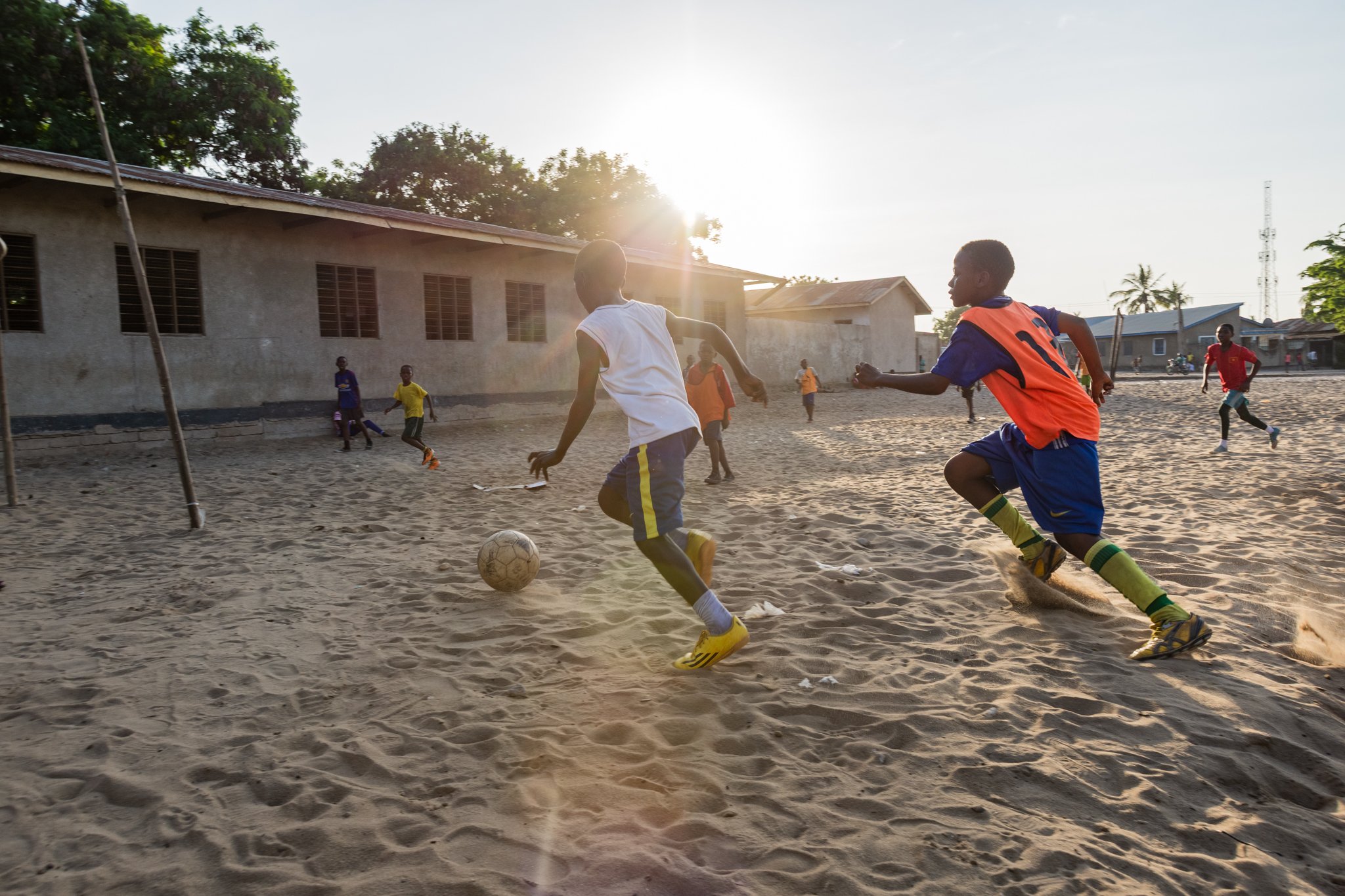 Children playing football on a sandy field near buildings with the sun setting in the background in Mbagala, Dar es Salaam.