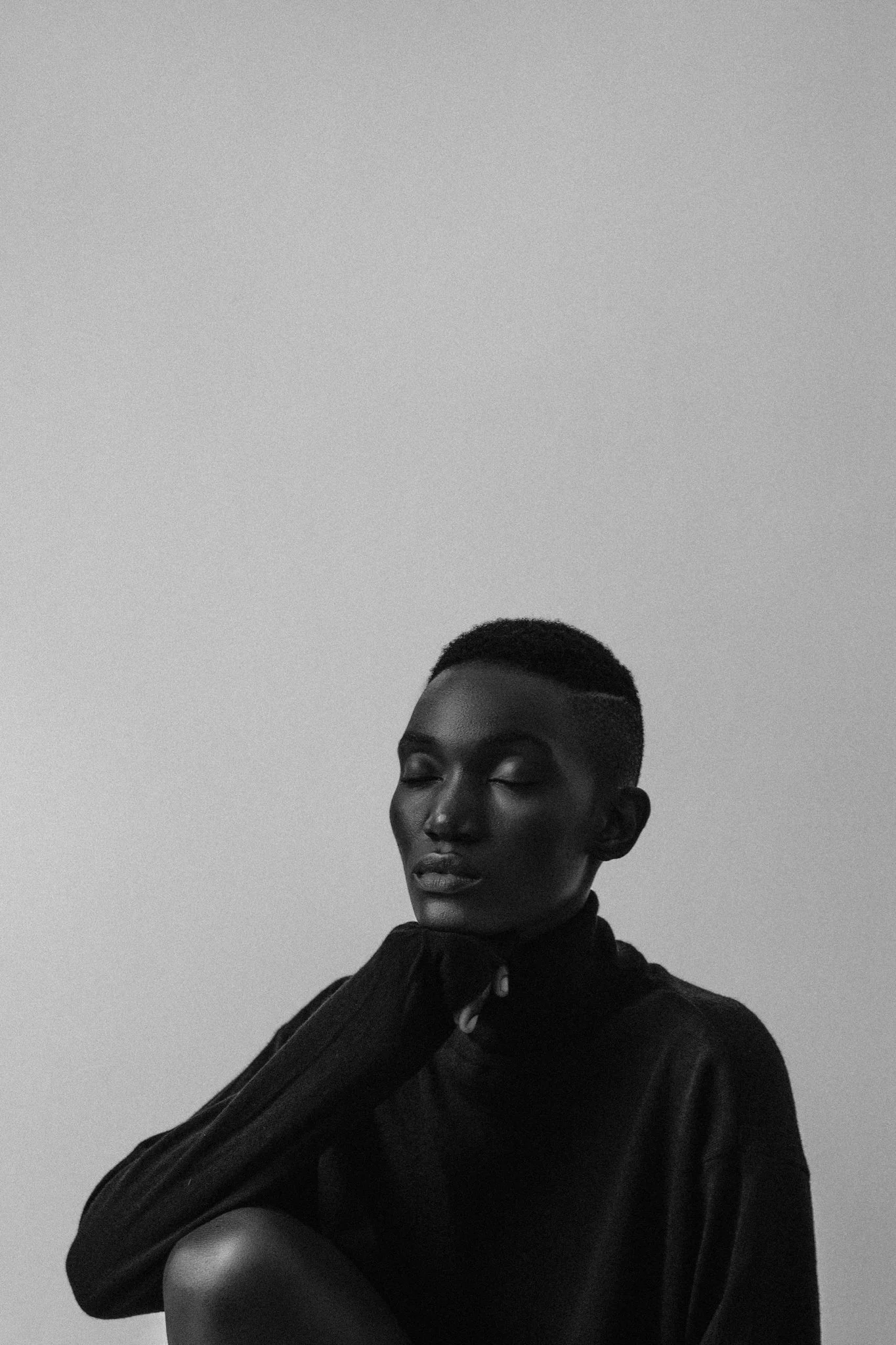 A high-contrast, black and white fine art photo of a female model with short hair and closed eyes, resting her head on her hand, wearing a dark, high-neck top against a plain background in Dar es Salaam, Tanzania.