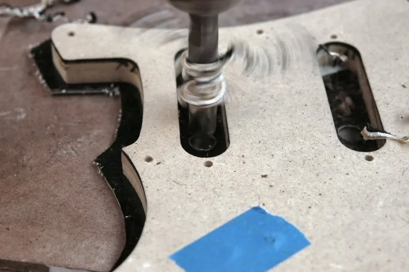 I make my own pickguards. Those available from the many manufacturers seem to vary considerably, even from the same supplier. This way, I can only get mad at my self. I take a piece of PG material, and trace the outline from a PG template, then cut it out on the band saw. My trusty Nikon was set on the wrong setting so you will have to imagine, tortoise shell plastic, MDF template, Sharpie marker, and my stubby fingers. I’ll use golf grip tape to double stick it to the template and step over to the drill press to drill the holes. Here the access for the pickup holes.