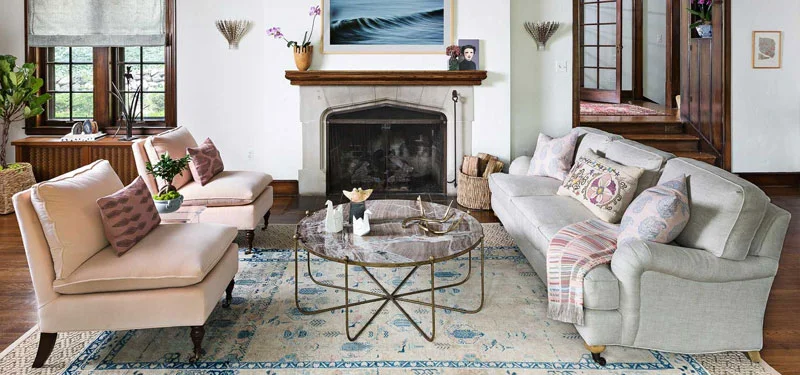 Picture of a sitting area designed by CLOTH & KIND in Ann Arbor, MI with white walls, dark wood floors, two light pink chairs, a white couch, blue and white patterned rug, and a granite coffee table. Text on the image reads "Ann Arbor Hills Tudor".