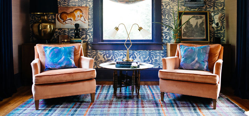 Picture of a sitting area designed by CLOTH & KIND in Ann Arbor, MI with light orange velvet chairs, a multicolored rug, blue palm wallpaper and gold lamps. Text on the image reads "Wilcox Modern Bungalow".