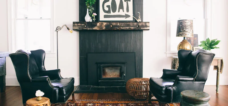 Picture of a living room designed by CLOTH & KIND in Ann Arbor, MI with a white walls, a black fireplace with a print that says "GOAT," black leather chairs, and a wooden table with a gold lamp. Text on the image reads "Saltbox on the Hill".