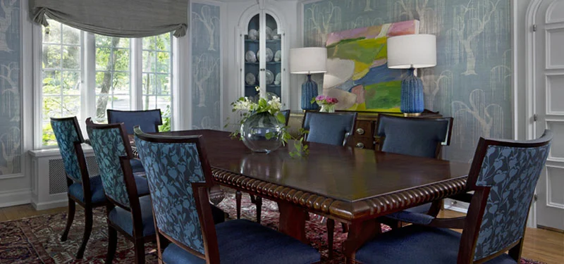 Picture of a dining room designed by CLOTH & KIND in Ann Arbor, MI with light blue wallpaper with trees, large dark wooden dining table, dining chairs covered in blue fabric, blue lamps and art. Text on the image reads "Ives Woods Tudor".