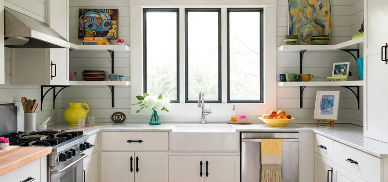 Picture of a kitchen designed by CLOTH & KIND in Ann Arbor, MI with light grey walls, large windows, white cabinetry, and colorful art and kitchen accessories. Text on the image reads "University Drive Bungalow".