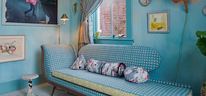 Picture of a gathering nook designed by CLOTH & KIND in Savannah, GA with blue textured walls, a large chaise lounge covered in light green and blue fabric, paint splattered floors, and art. Text on the image reads "Southern Style Now Showhouse".