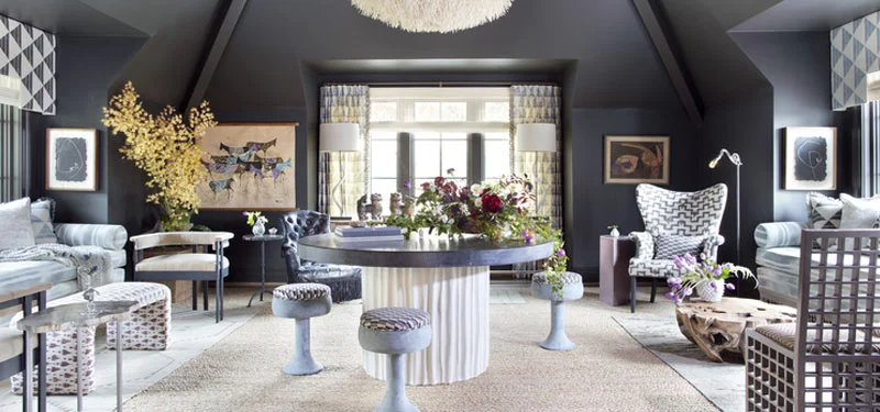 Picture of a room designed by CLOTH & KIND in Ann Arbor, MI with a large circular table, purple patterned stools, two sitting areas with patterned couches and chairs, cream light fixture, and colorful art. Text on the image reads "Holiday Showhouse".