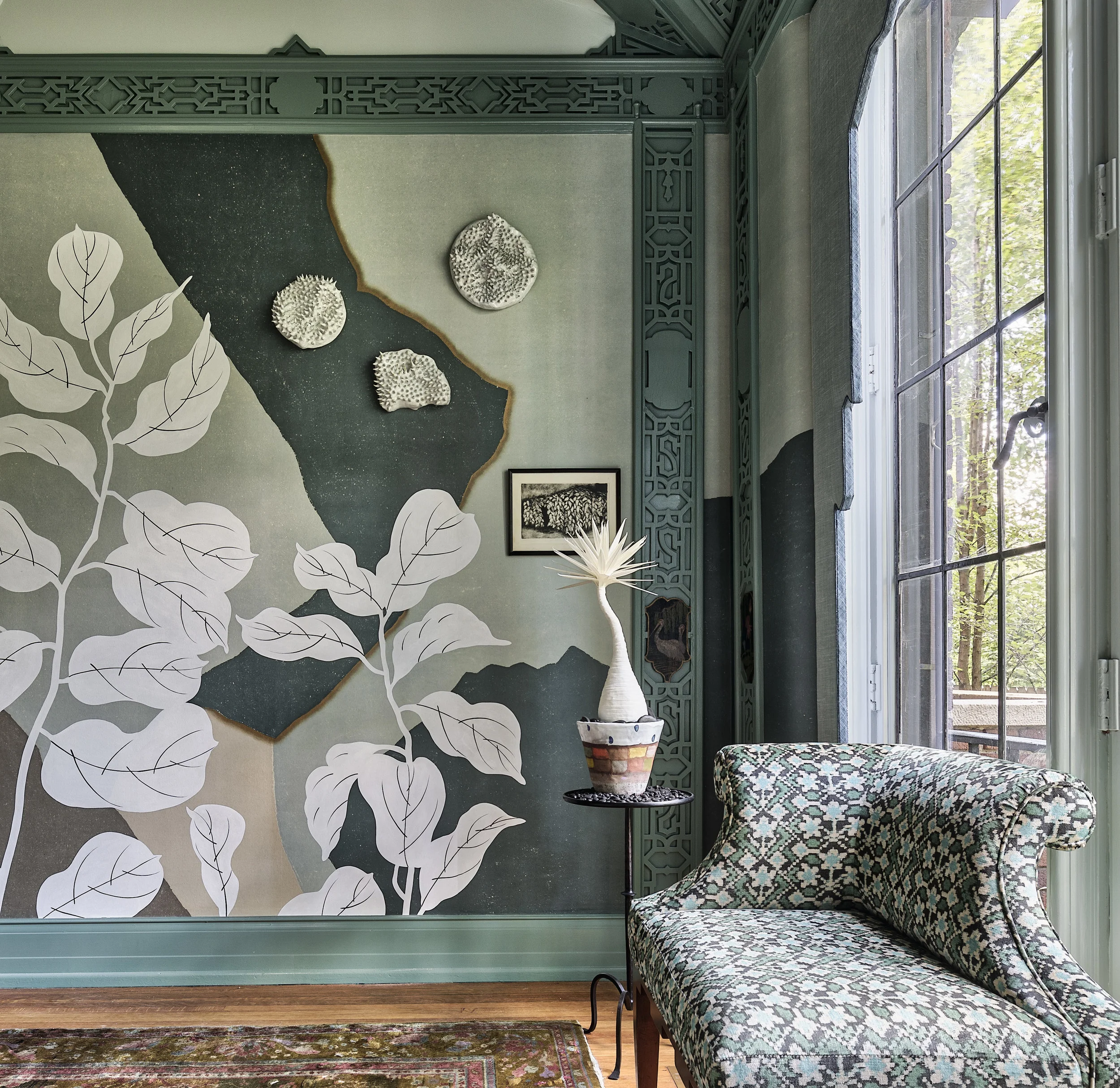 Picture of a corner in a breakfast nook designed by CLOTH & KIND in Ann Arbor, MI with a layered wall mural including a white plant, a green and blue patterned chaise, and a textured rug. Text on the image reads "Charles T. Fisher Mansion Showcase".