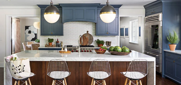 Picture of a kitchen designed by CLOTH & KIND in Ann Arbor, MI with a white subway backsplash, navy cabinetry, wooden island with white granite countertop, and silver bar chairs with white cushions. Text on the image reads "Highland Tudor Gut Rehab".