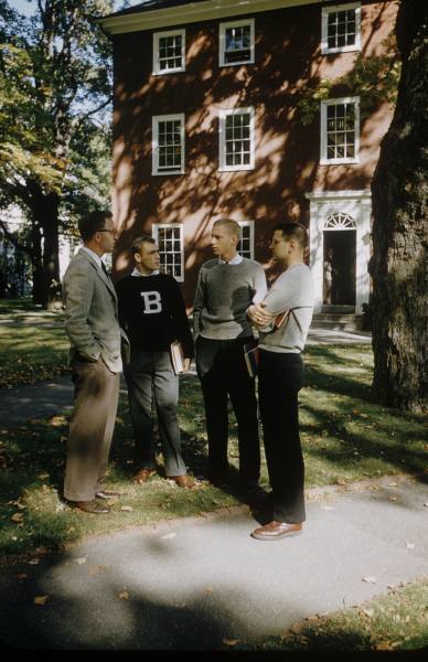 A group of Bowdoin College men stand around talking outside a dormitory. One of them is wearing a big B letter sweater and gray slacks.