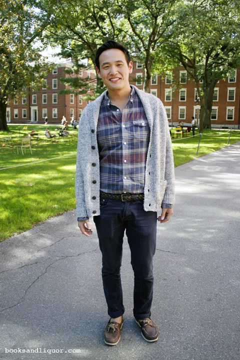 Harvard University student wearing a plaid shirt under a gray wool cardigan and over blue jeans with a woven brown leather belt. He is wearing brown boat shoes