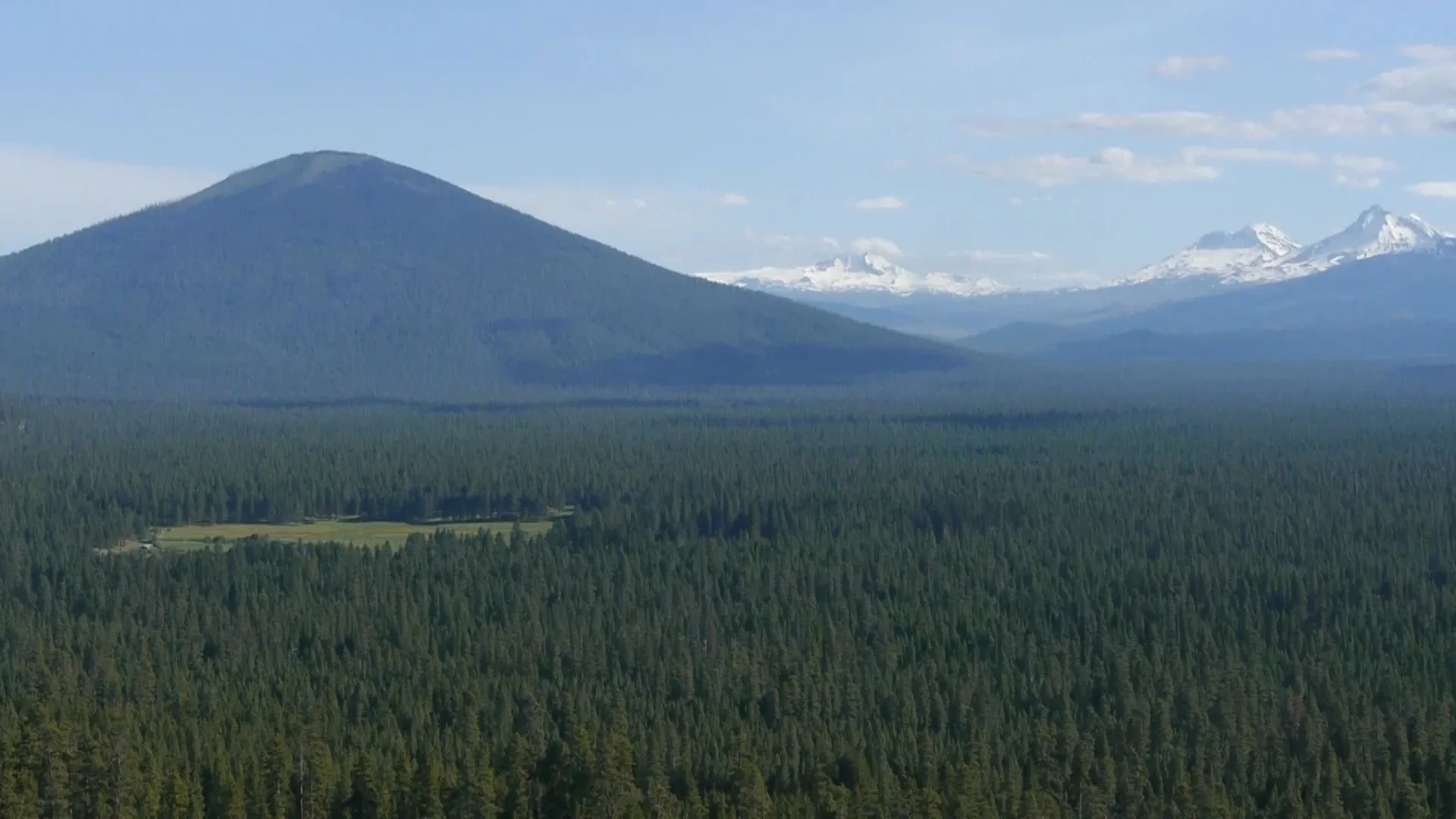 View of Black Butte with the meadow of House on Metolius as a sliver of light green in the sea of Ponderosa pine. The North, Middle and South Sister are visible on the horizon. Photo by Marta Van Patten.