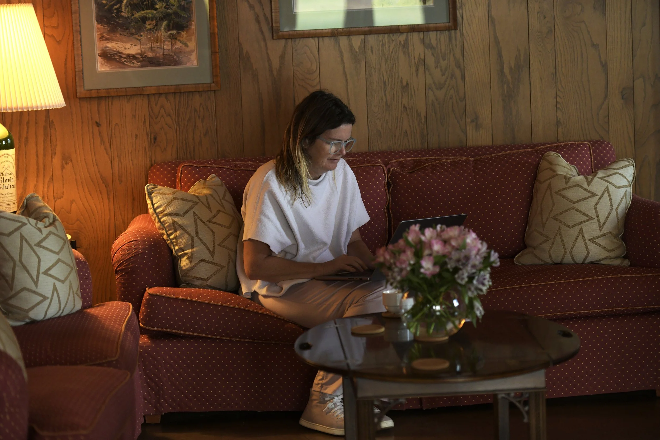 A woman with glasses, wearing a white t-shirt, sitting on a red patterned couch with plaid pillows, working on a laptop. There is a dark wood coffee table with a pink flower arrangement in a glass vase in front of her. The room has a wooden wall with
