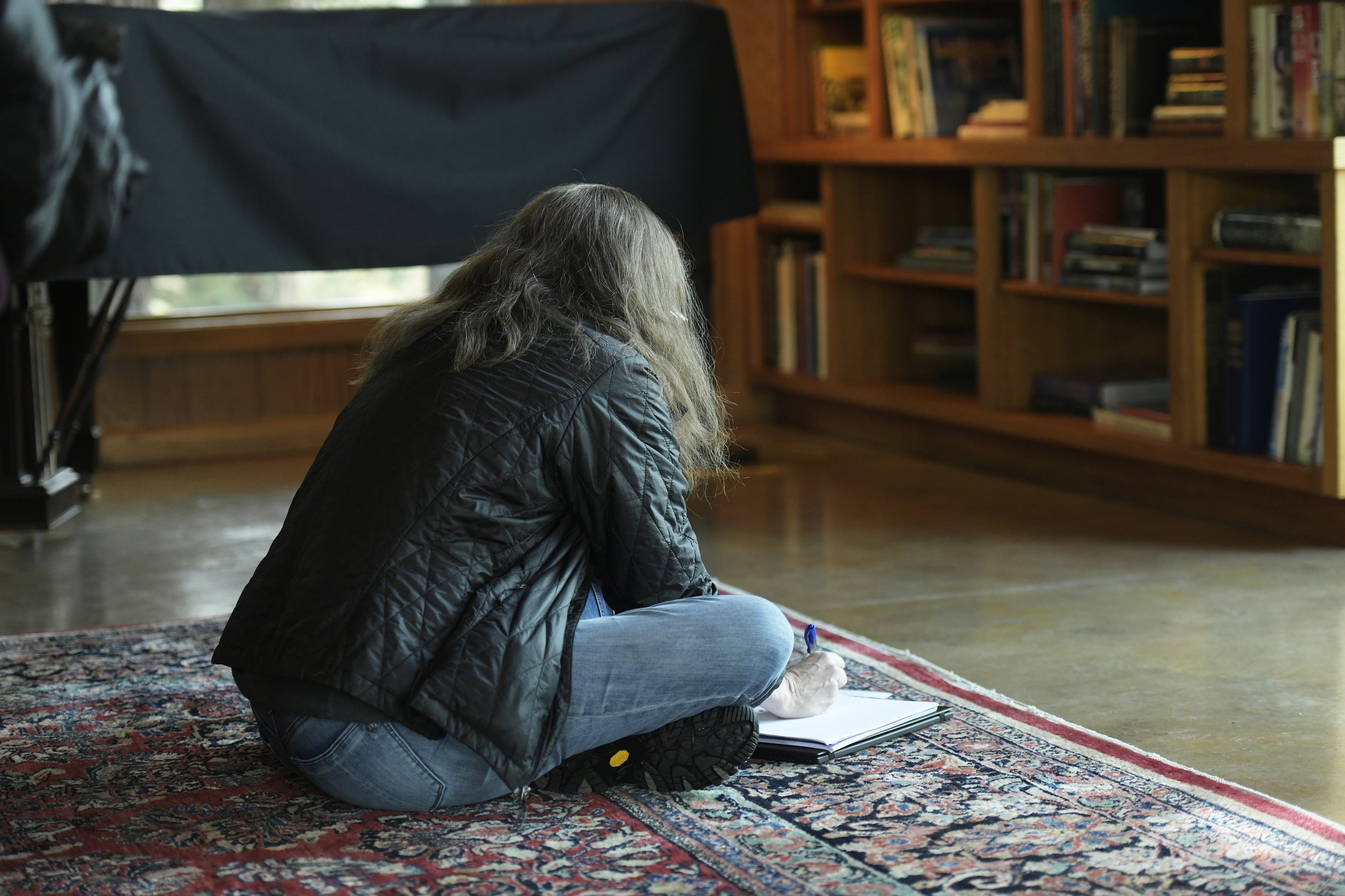 A person with long hair wearing a black quilted jacket and jeans sitting on a colorful Persian rug, writing in a notebook, with a bookshelf in the background.