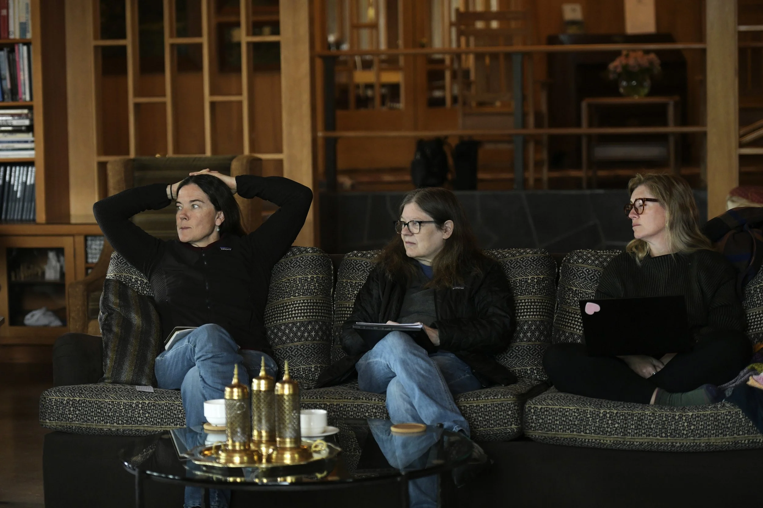 Three women sitting on a patterned sofa in a wooden room. The woman on the left has dark hair, is wearing a black jacket, and has her hands on her head. The woman in the middle has glasses, brown hair, and is holding a notepad. The woman on the right
