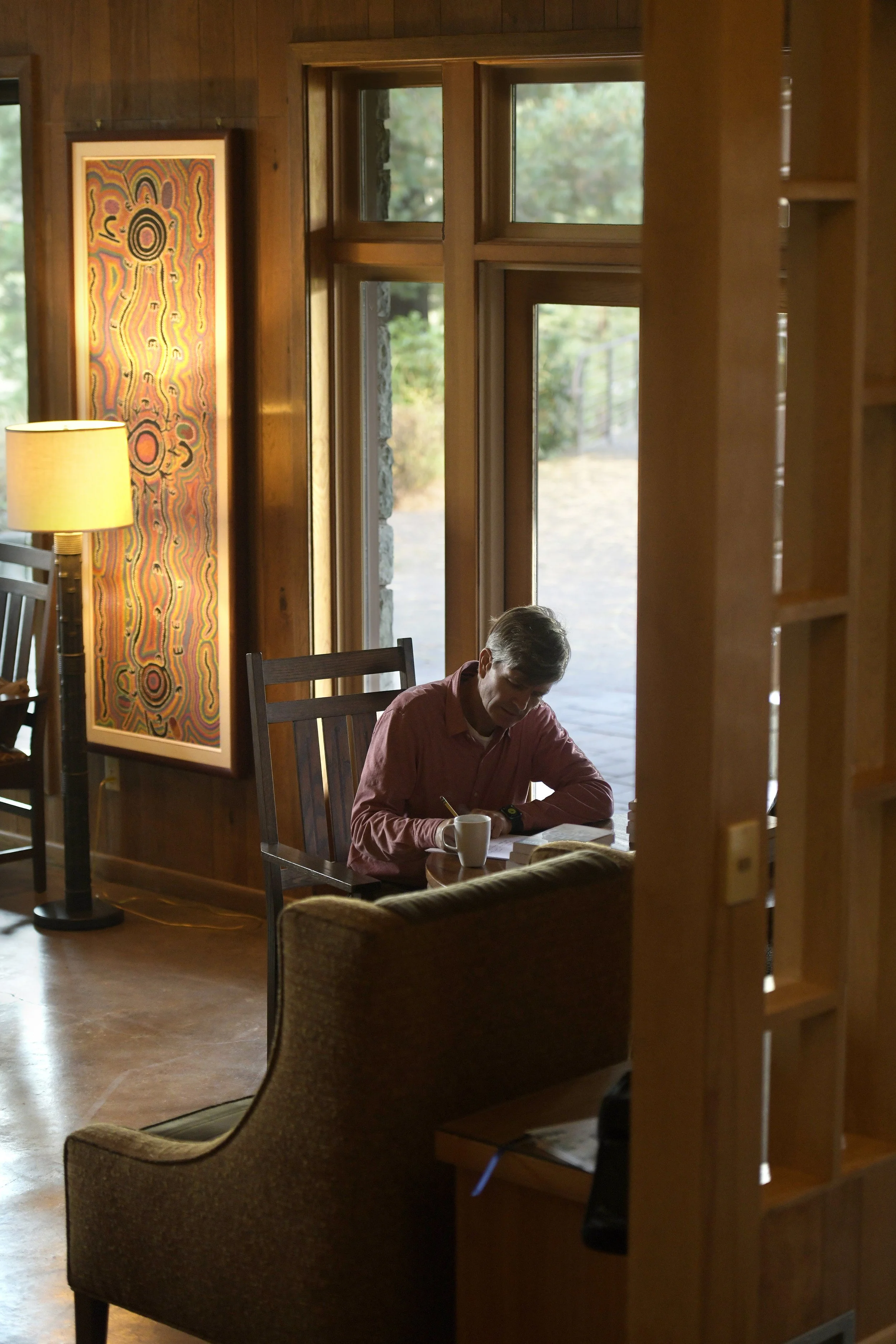 A man sitting at a wooden table in a cozy, wood-paneled room, reading and writing in a notebook with a coffee mug nearby. Large windows overlook a natural outdoor setting, and a colorful abstract painting hangs on the wall.