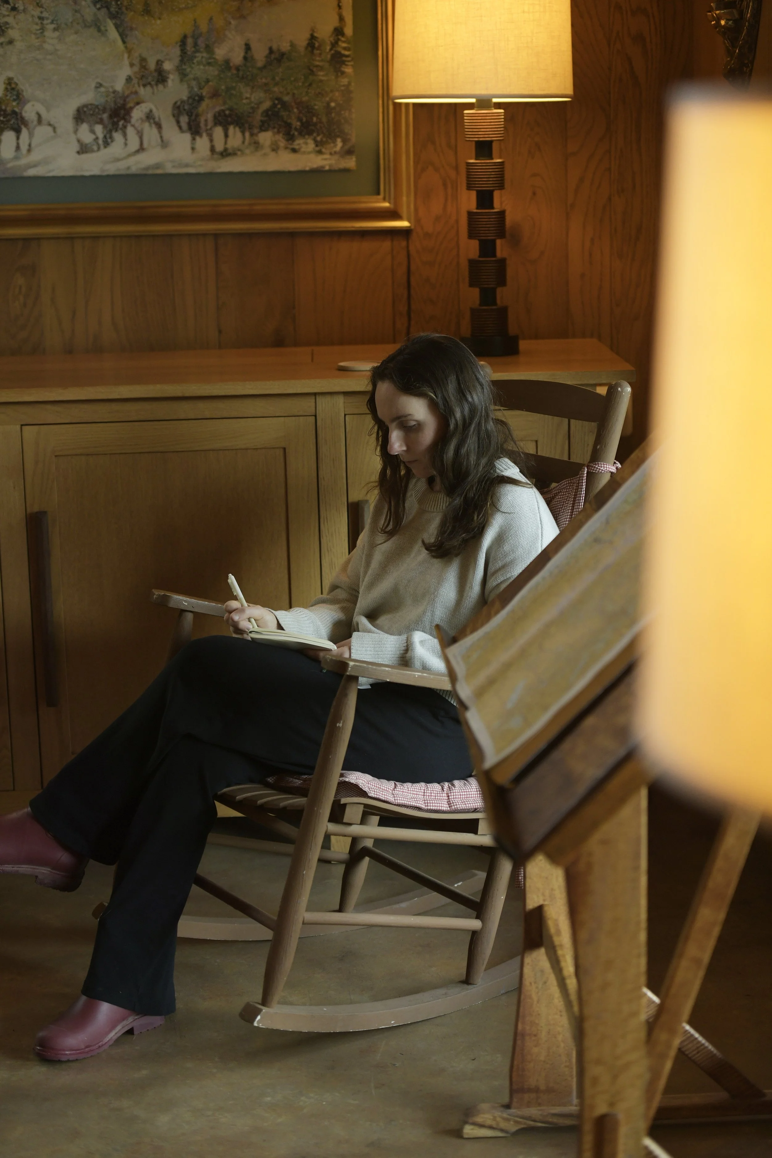 A young woman with long brown hair sitting in a wooden rocking chair, writing in a notebook. She is in a cozy, wood-paneled room with a painting of animals and a lit table lamp nearby.