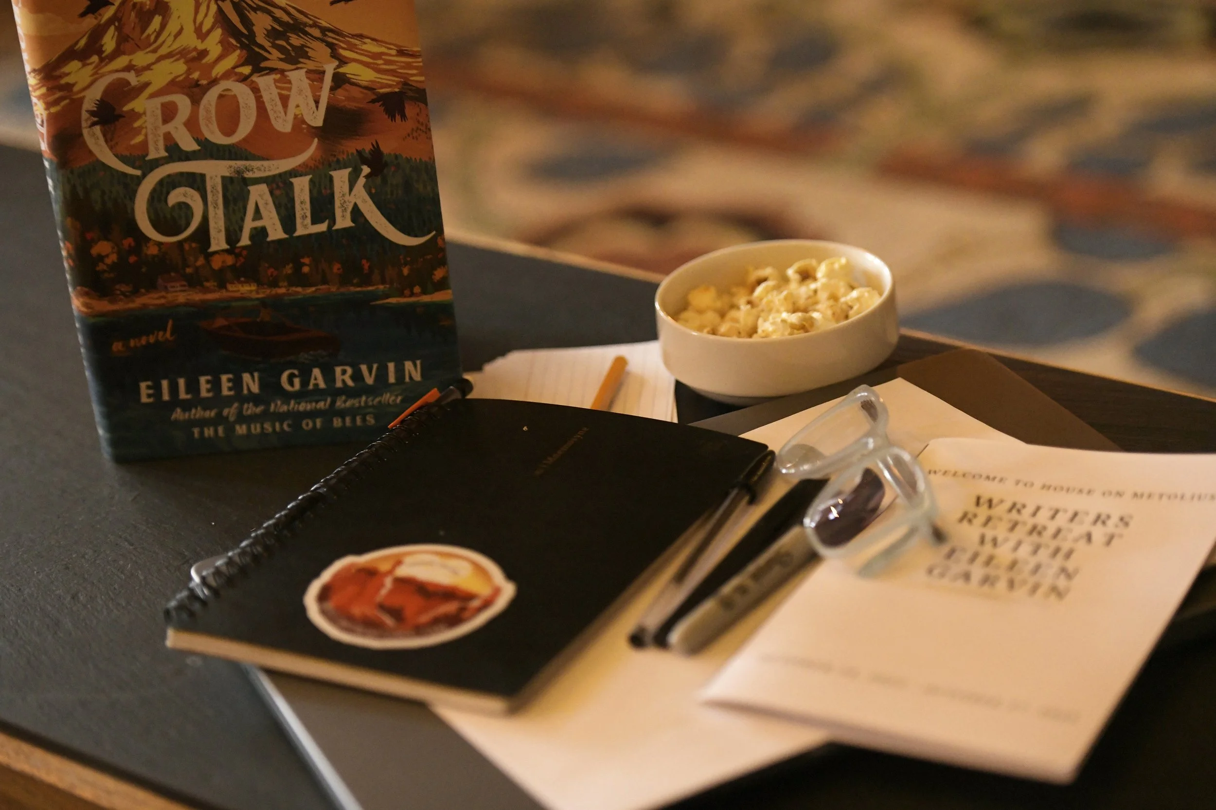 A book titled 'Crow Talk' by Eileen Garvin on a table, with a bowl of popcorn, a notebook, a pen, a pair of glasses, and a printed sheet of paper with the title 'Writers Retreat with Eileen Garvin' on it.