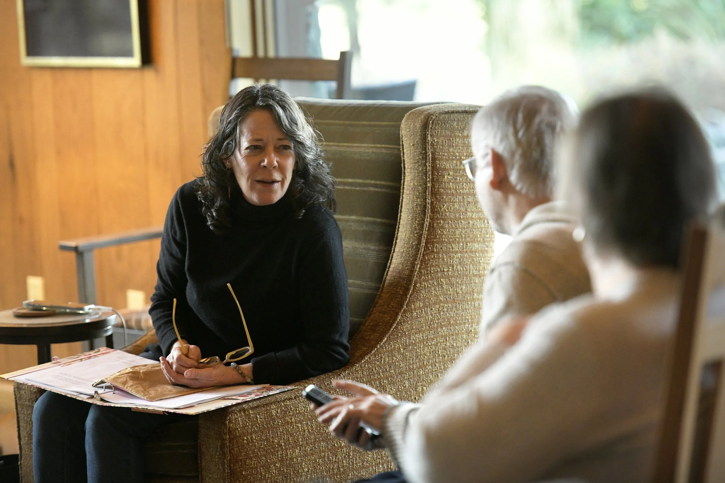 Three people sitting on a couch in a cozy living room, having a conversation. One woman with black hair and a black sweater appears to be talking, while an older woman with glasses and a beige sweater and an older man with white hair and glasses listen. A side table with a phone and newspapers is nearby, and sunlight filters through the windows.