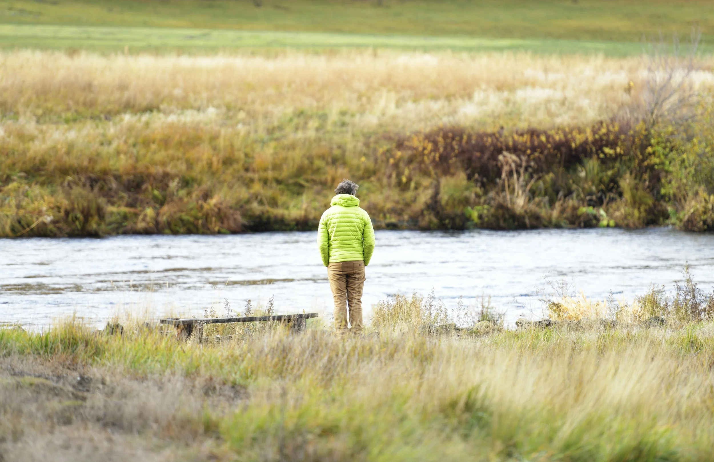 Person wearing a bright green jacket standing by a river with autumn foliage in the background.
