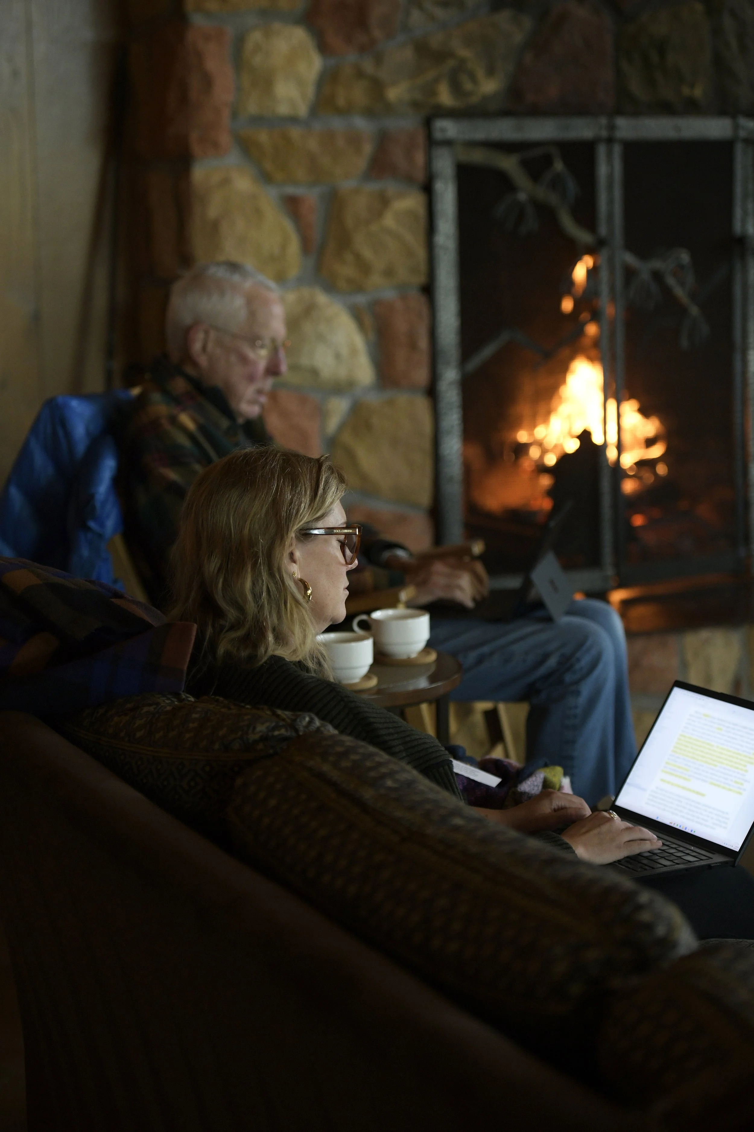 Two people sitting in front of a fireplace with a fire burning, working on laptops, with cups on a small table beside them.