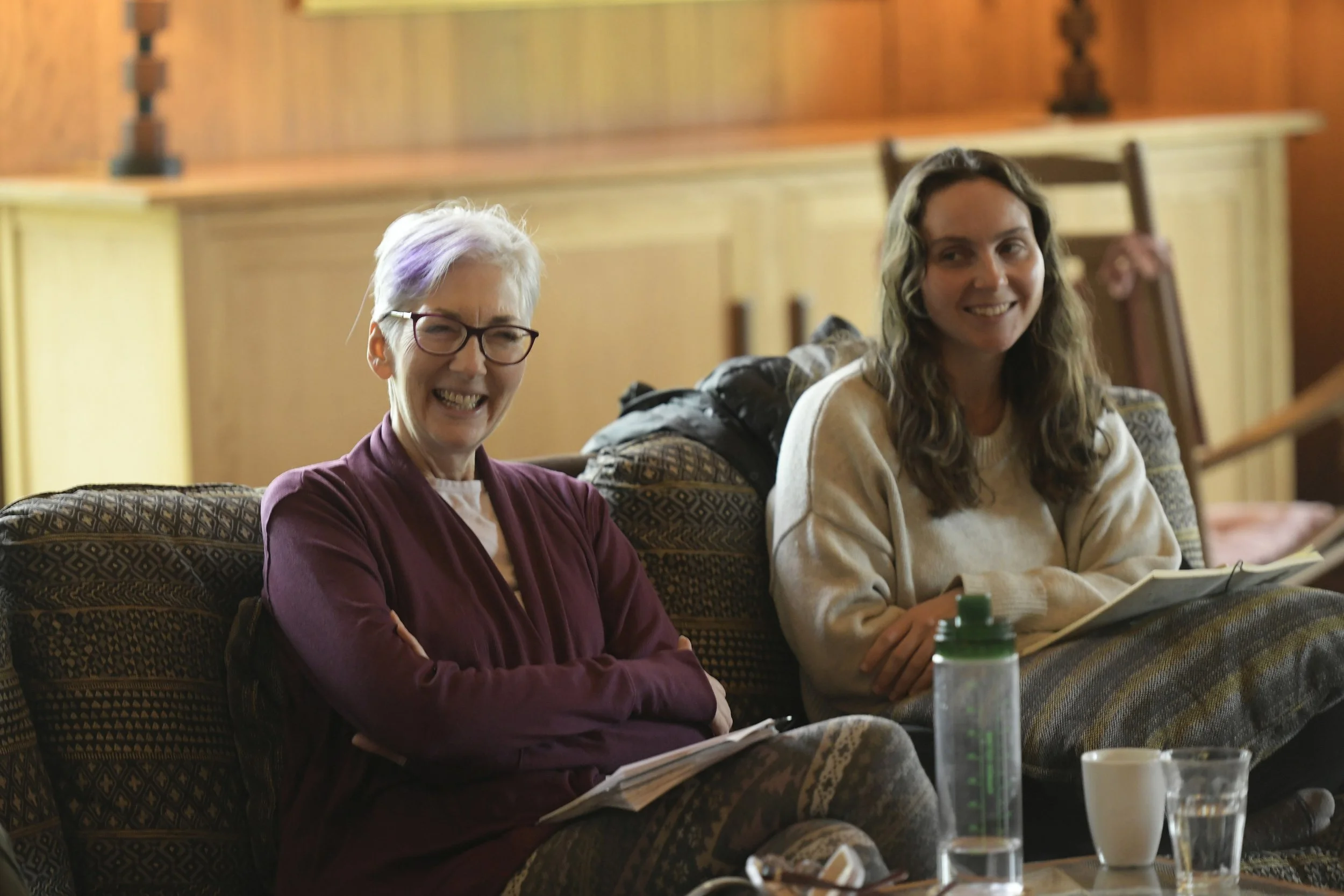 Two women sitting on a patterned couch, smiling and talking. The older woman has short gray hair, glasses, and a maroon jacket. The younger woman has long wavy brown hair and a beige sweater. There are notebooks, a green water bottle, a coffee cup, a