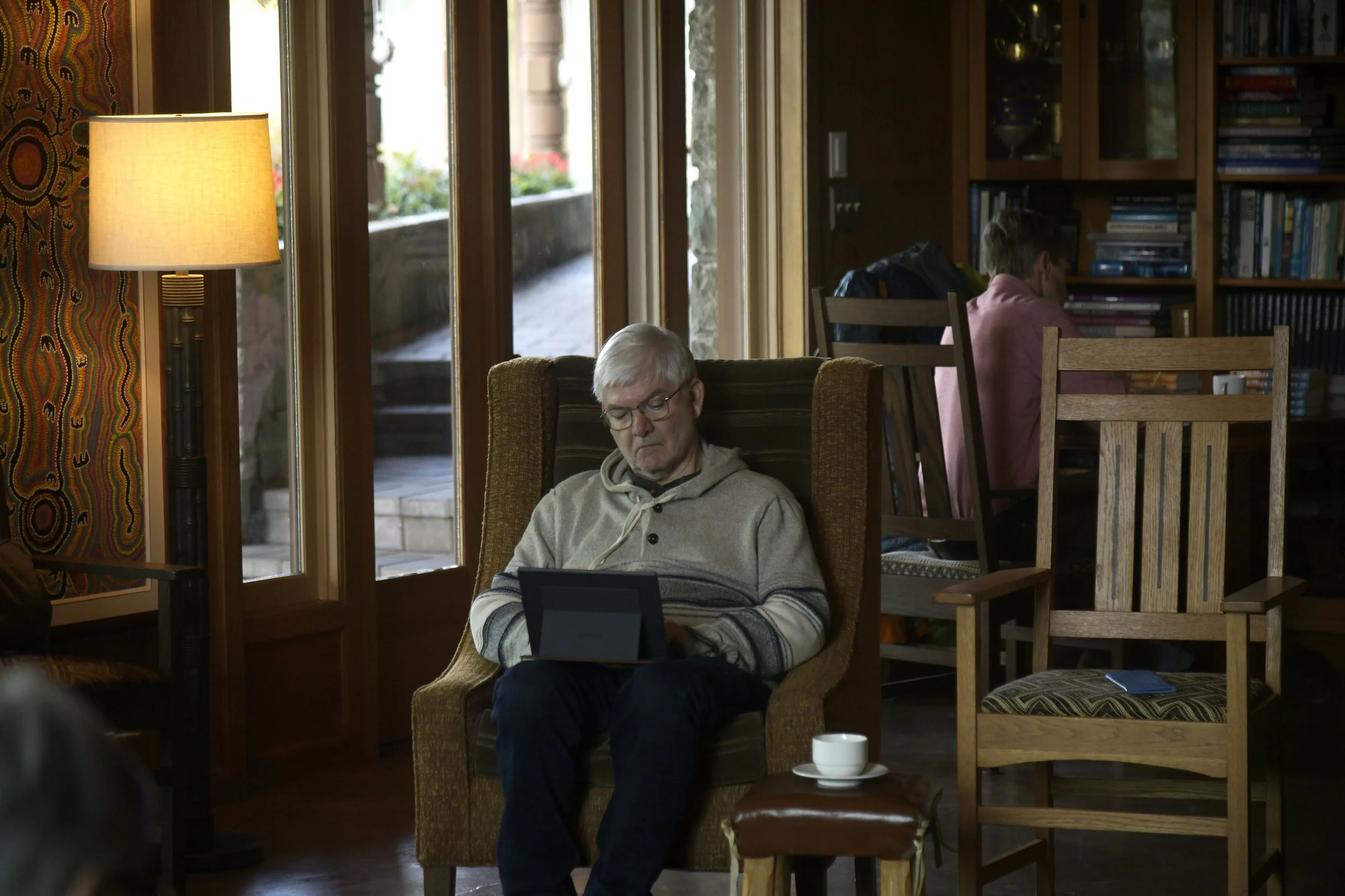 An elderly man with glasses and gray hair sitting in a brown armchair, reading or looking at a tablet, in a cozy room with wooden furniture, a lamp, and large windows letting in natural light, next to a small table with a white cup.