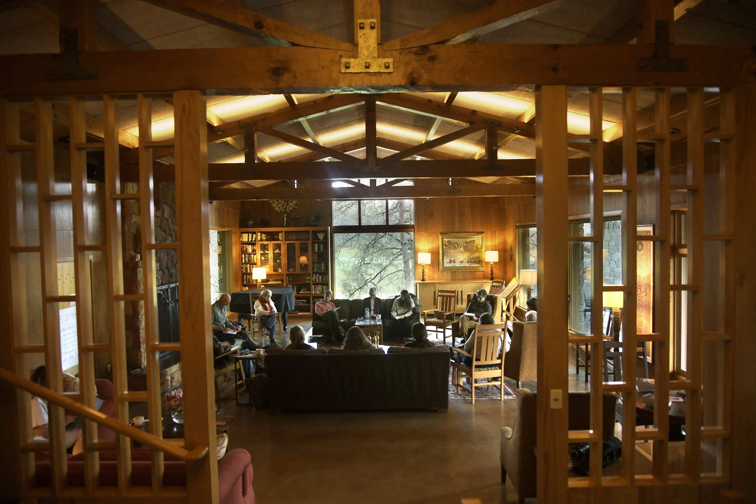 A cozy indoor gathering or meeting room with wooden walls and ceiling. Several people are seated on chairs and sofas, engaged in conversation. Large windows overlook a natural landscape, and the room is warmly lit with lamps and candles. A bookshelf with books and decorative items is against the back wall.