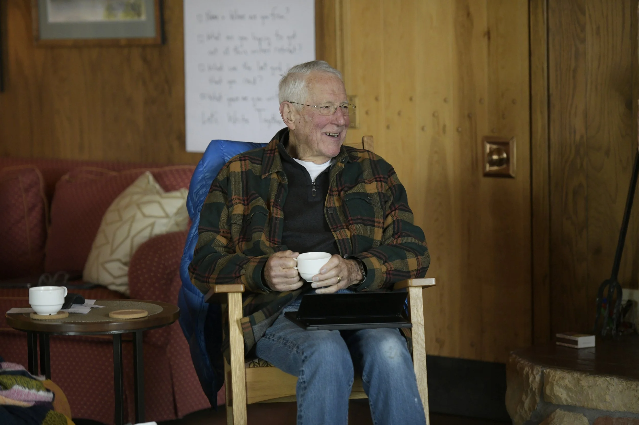 An elderly man with white hair and glasses sitting on a wooden chair, smiling and holding a white mug. He is wearing a plaid shirt, black sweater and blue jeans. There is a small table with a white cup and dishes next to him, a sofa with pillows in t