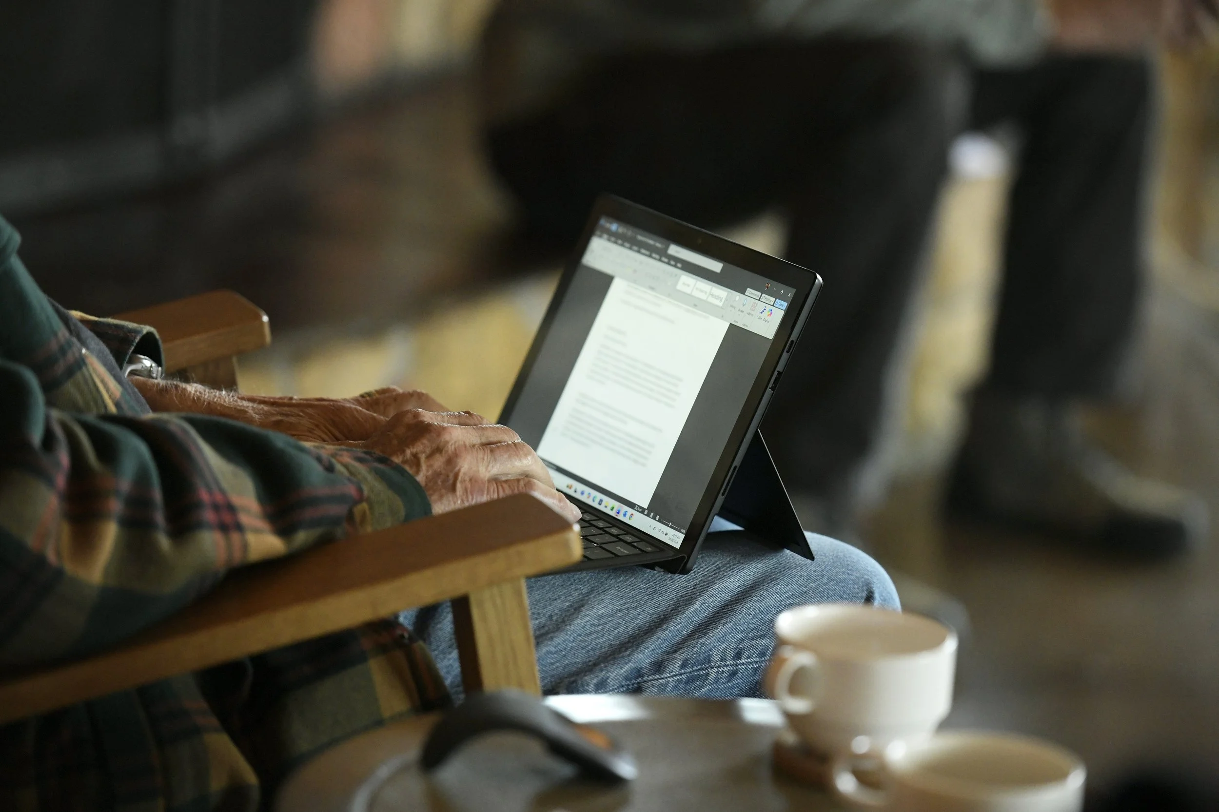 Person using a tablet on their lap, with a focus on their hands and plaid shirt, seated in a wooden chair in a cozy setting with a cup of coffee on a nearby table.
