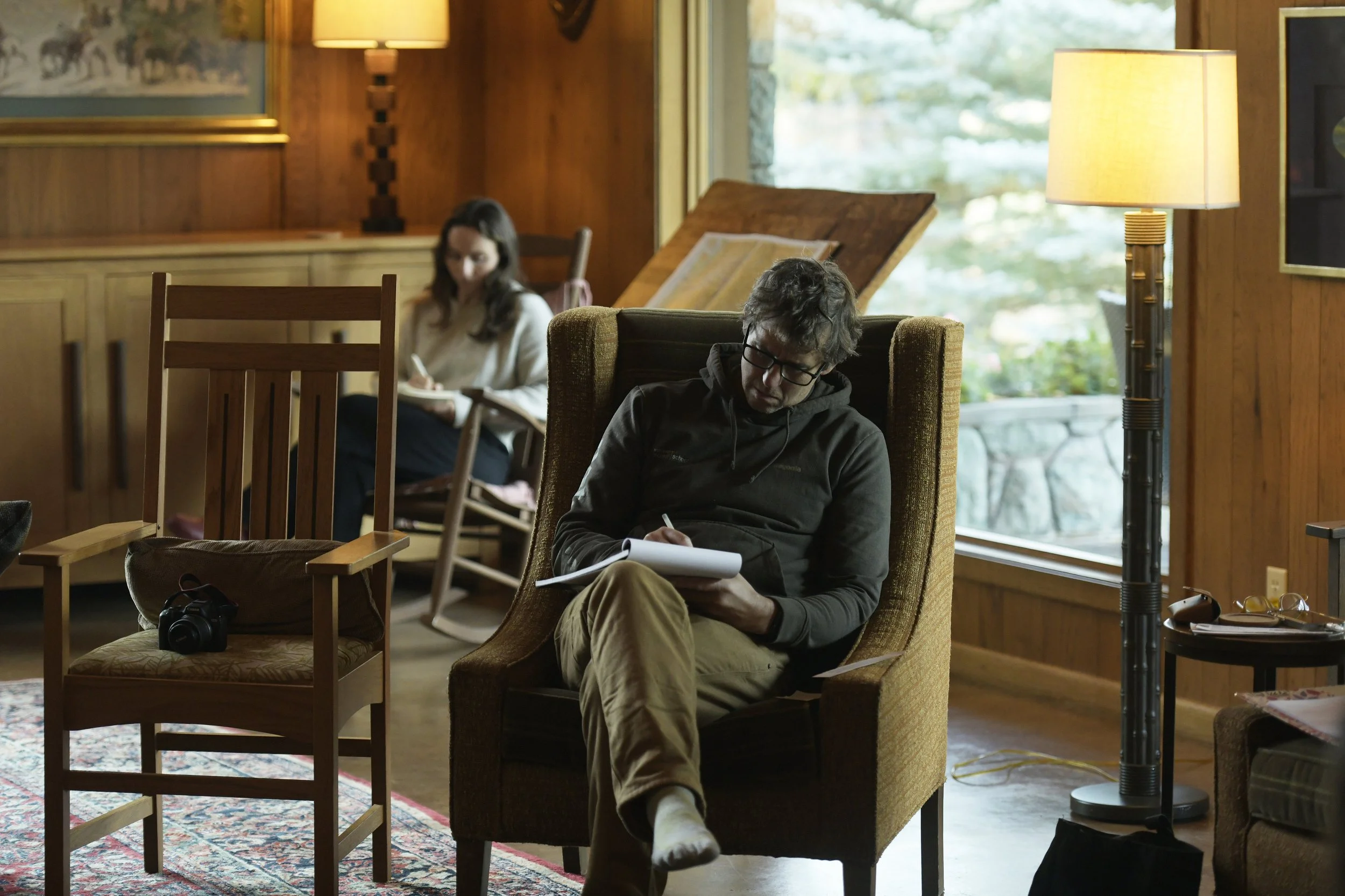 Two people are in a cozy living room with wood-paneled walls, engaged in writing or sketching. One person, with glasses and dark hair, is sitting in an armchair near a large window, focused on his notebook. The other person, with long dark hair, is s