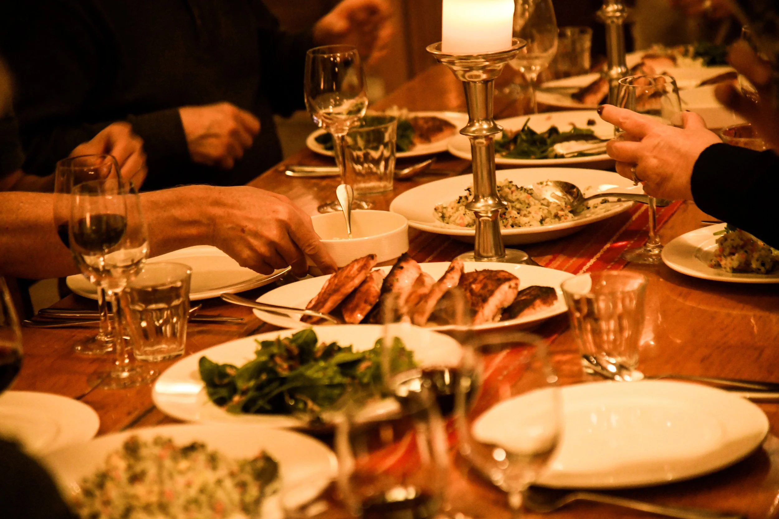 People gathered around a warmly lit dining table sharing a meal, with plates of grilled meat, salads, wine glasses, and a lit candle in the center.