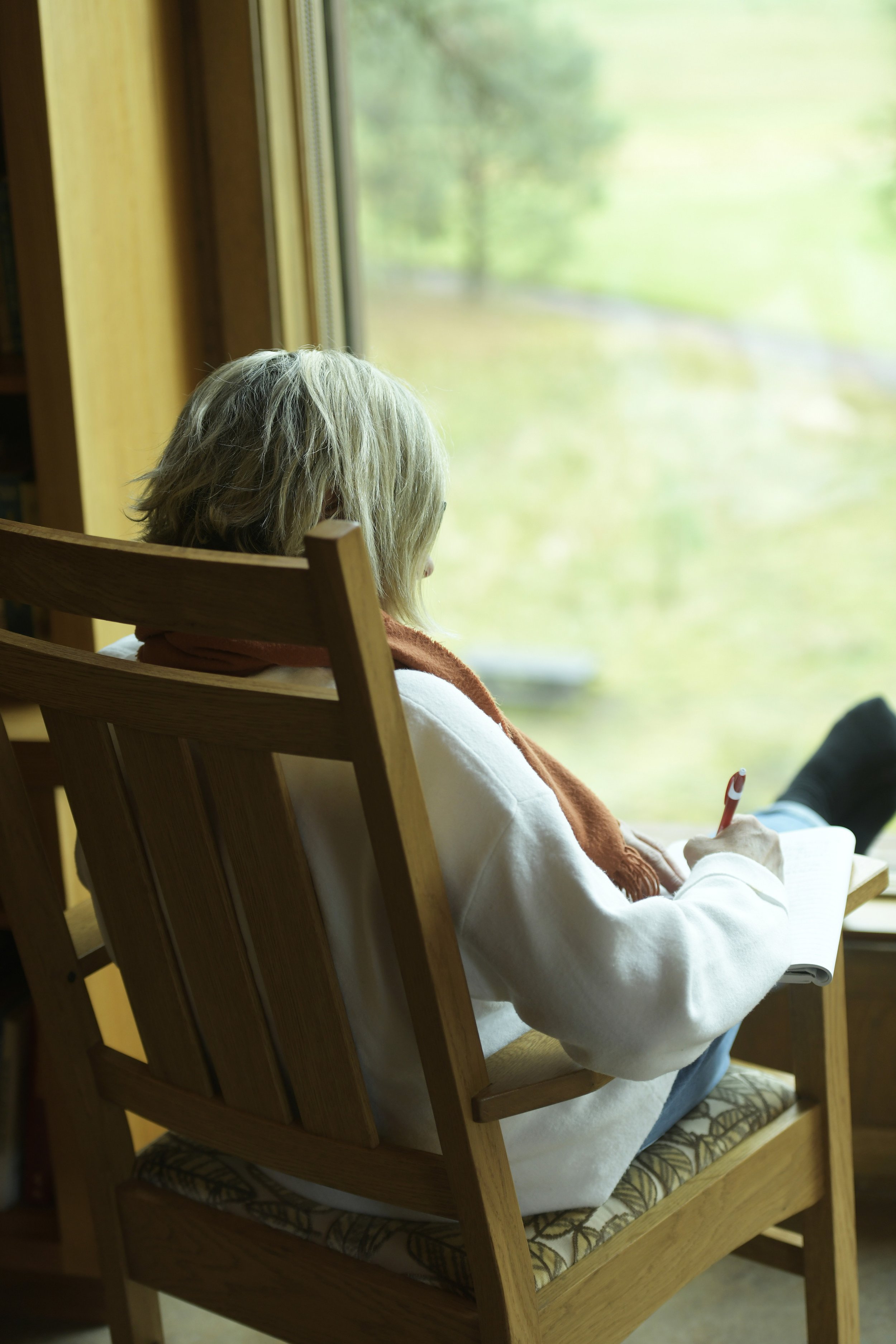A woman sitting in a wooden chair by a window, writing in a notebook with a pen, with a scenic view of green trees outside.