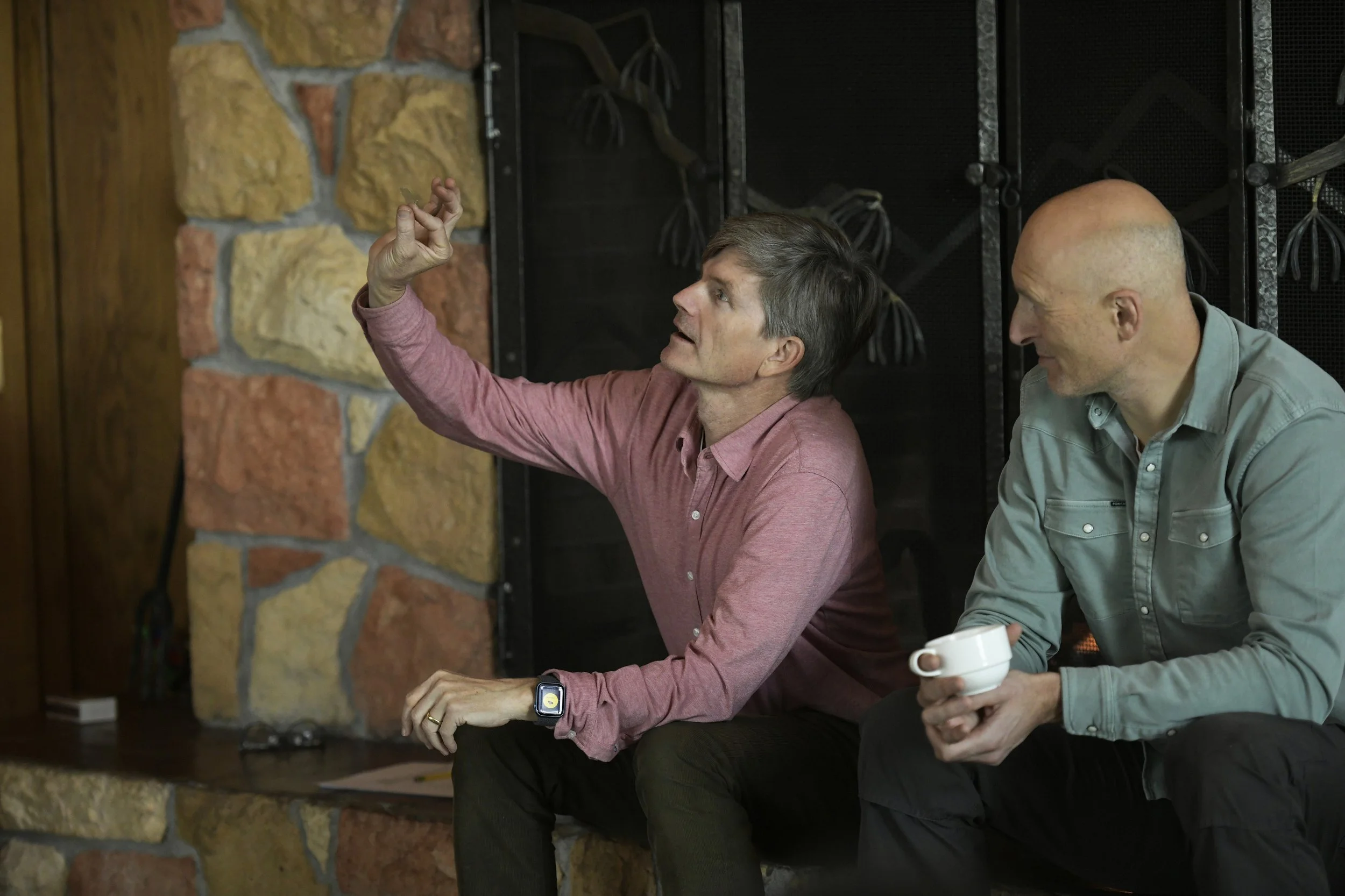 Two men sitting in front of a stone fireplace, one with gray hair and wearing a pink shirt making a gesture with his hand, the other bald and holding a white mug, engaging in conversation.
