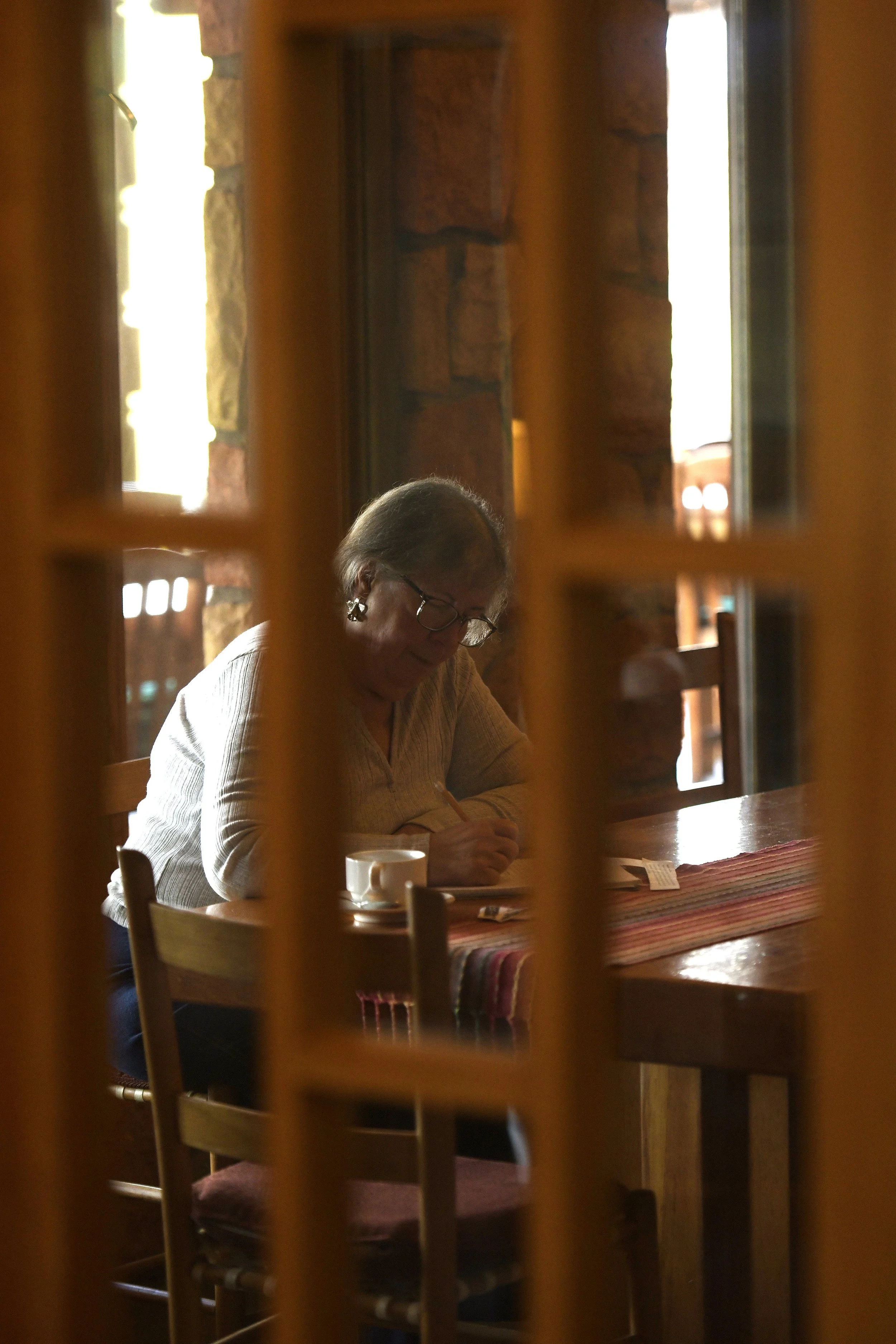 A woman with gray hair, glasses, and earrings sitting at a wooden table, writing or reading, with a cup in front of her, seen through a wooden lattice in a warmly lit room with stone walls.