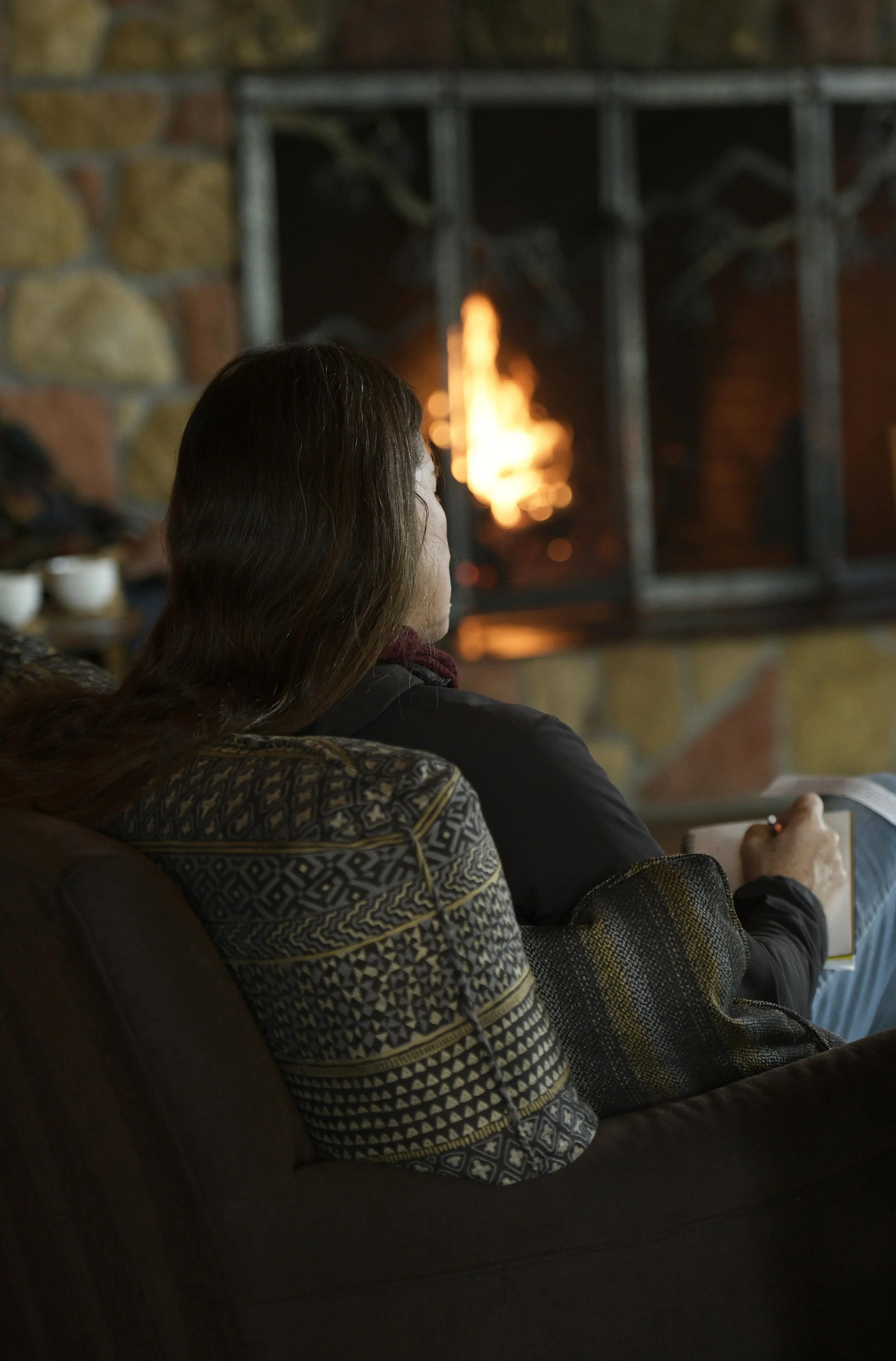 Woman sitting on a couch by a fireplace, reading a notebook.