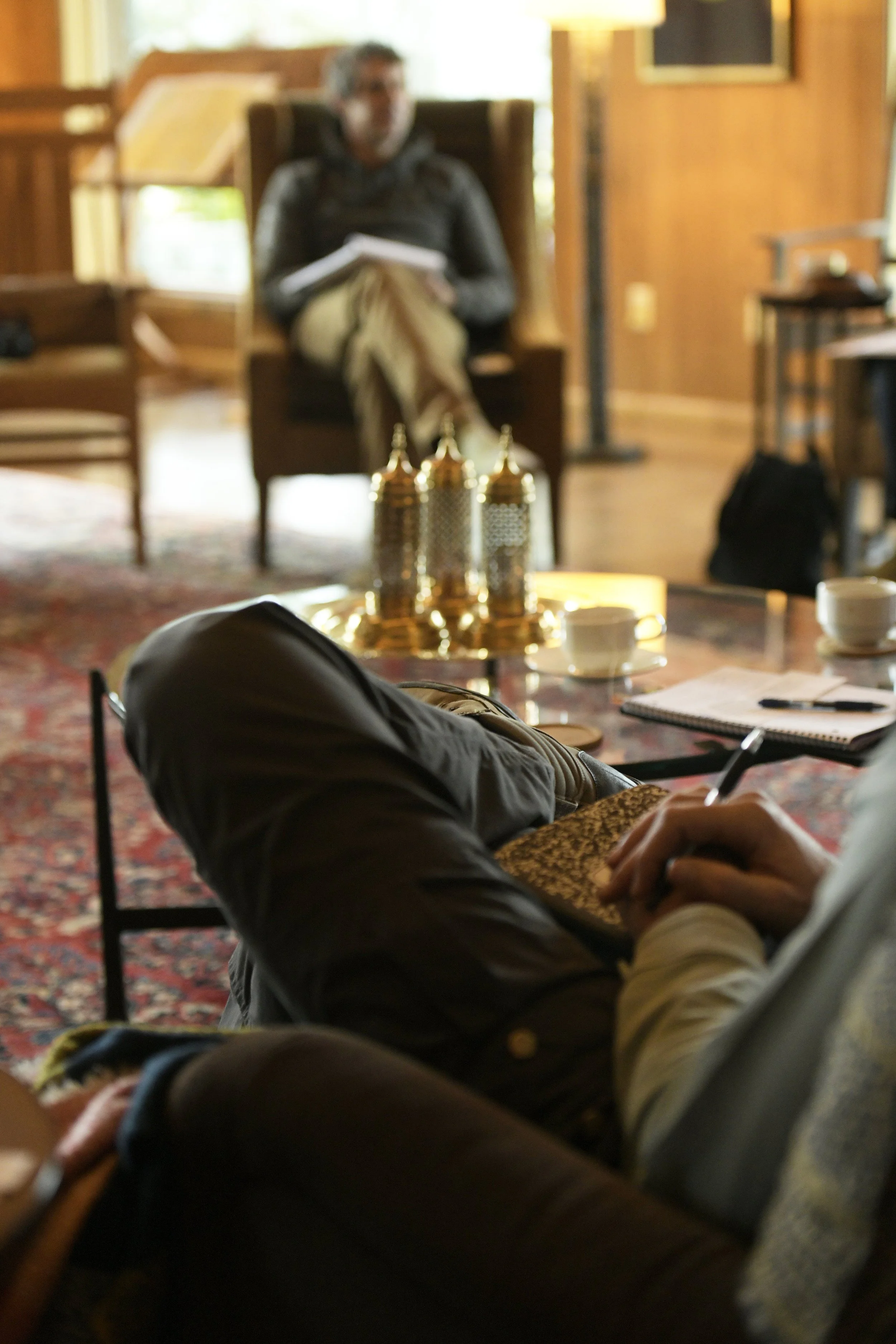 A group of people sitting in a cozy room with wooden walls, some taking notes. In the foreground, a person is writing in a notebook. In the background, a woman is seated on a chair holding a notebook, with a table of decorative gold items and cups in