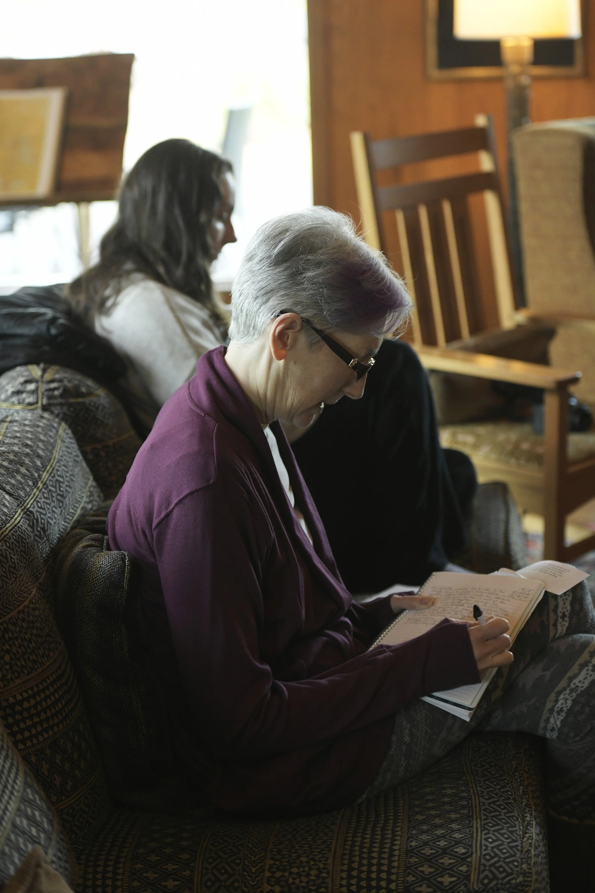 Two women sitting on a patterned couch in a cozy living room. One older woman with short gray hair, glasses, and a maroon cardigan writing in a notebook. A younger woman with long dark hair sitting behind her, looking down.