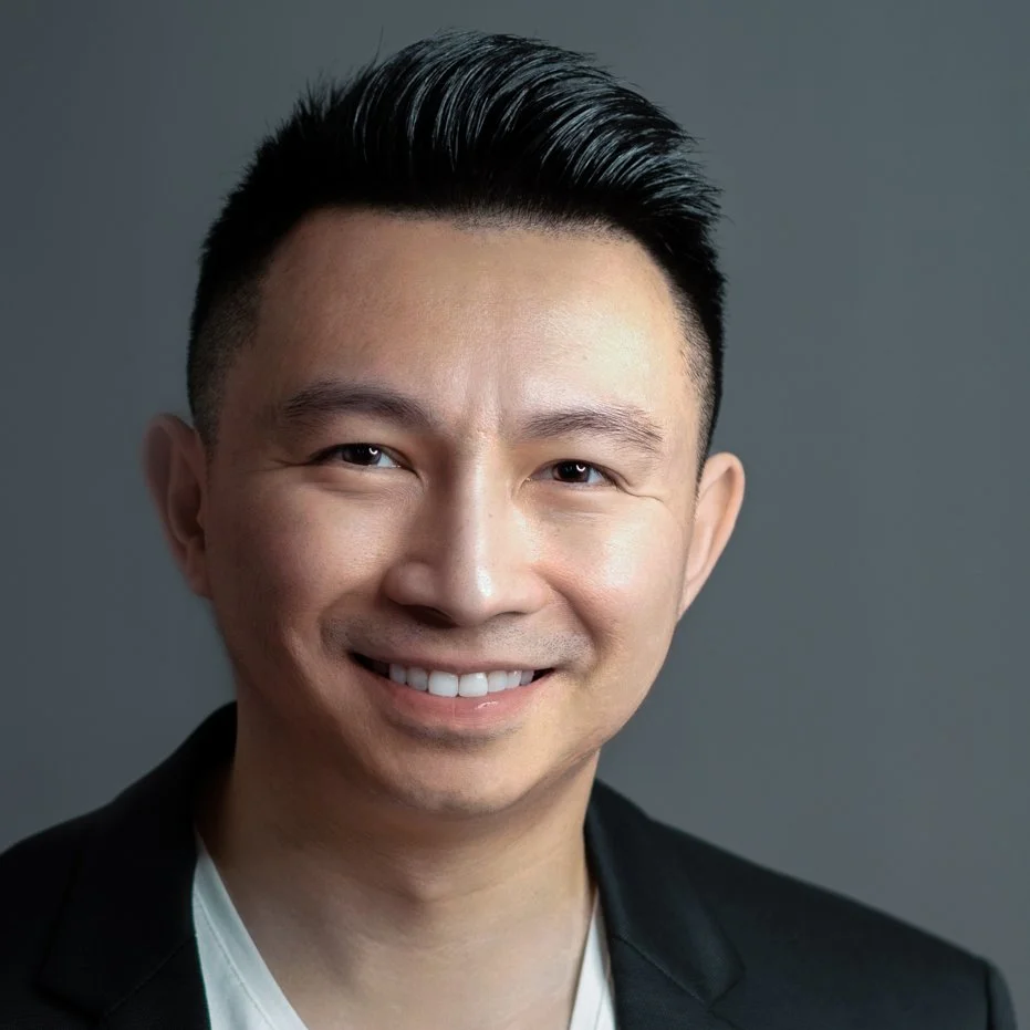 Smiling man with short hair, wearing a black blazer and white shirt, against a gray background.