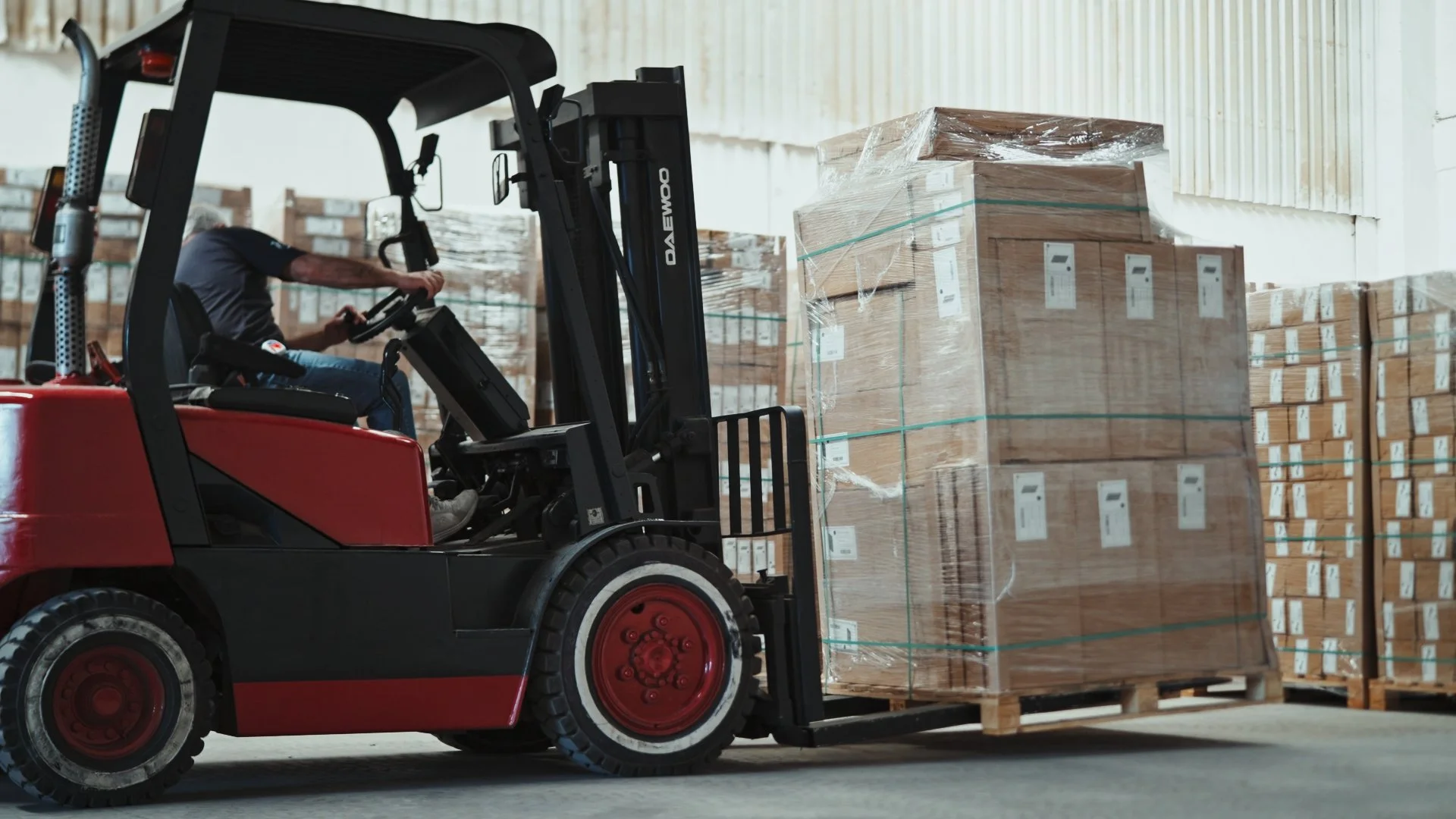 A person operating a red forklift in a warehouse, lifting a pallet loaded with cardboard boxes onto a rack.