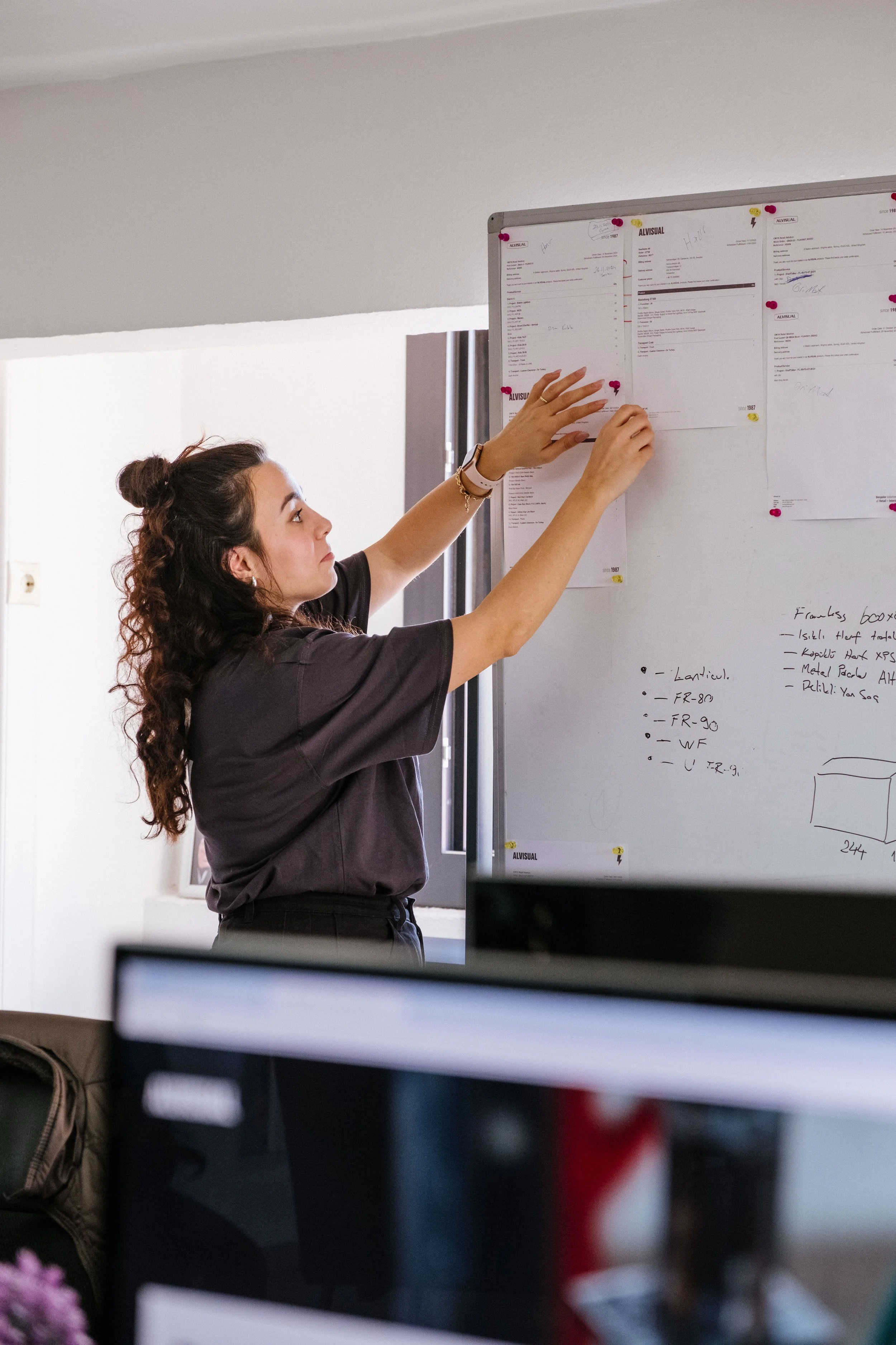 A woman with long curly hair hanging loose, wearing a dark t-shirt, is pinning papers to a whiteboard filled with documents and handwritten notes in an office.
