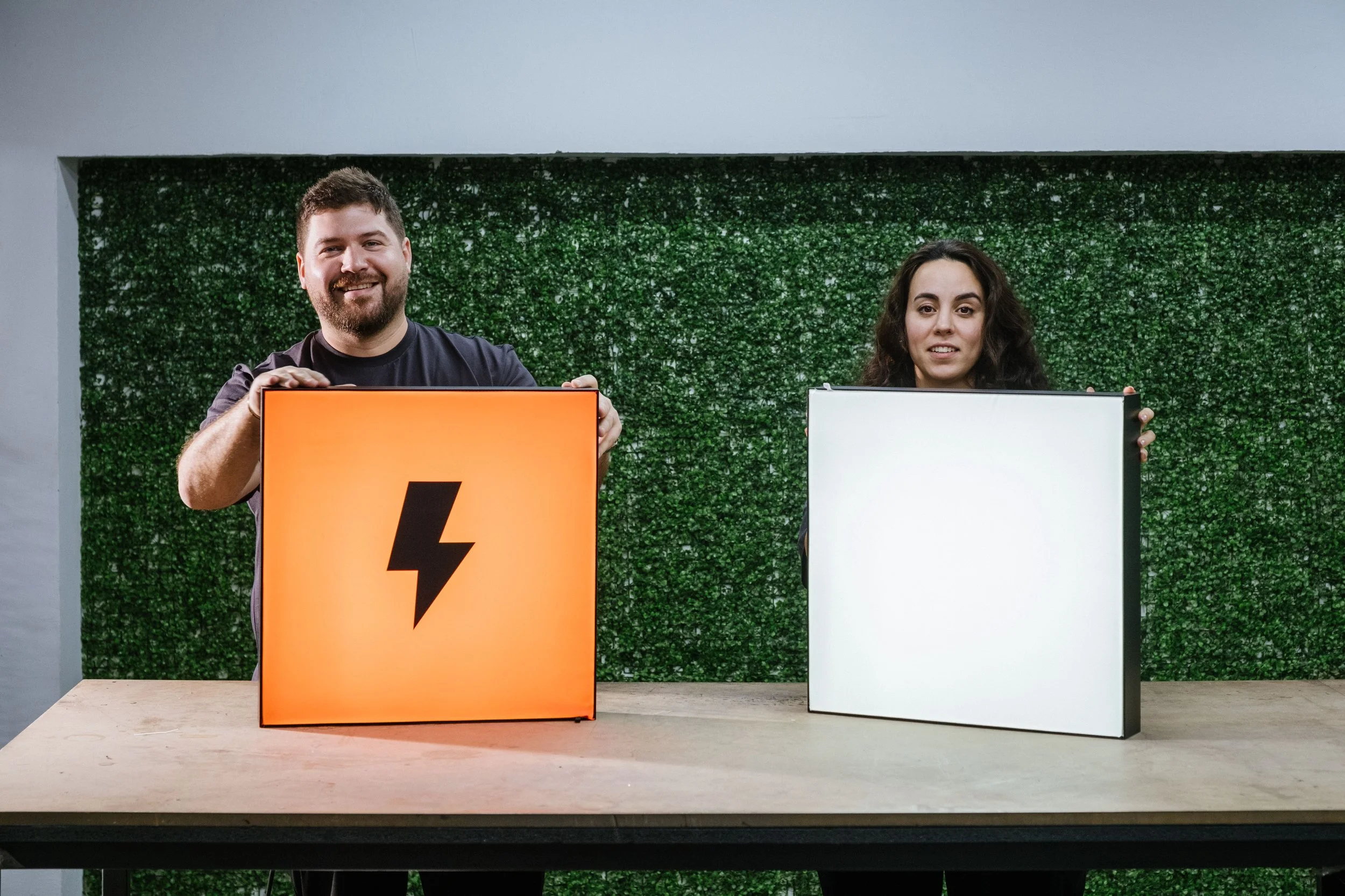 Two people are standing behind a table, holding illuminated square signs with a lightning bolt symbol on the left and a blank white square on the right, in front of a green textured wall.