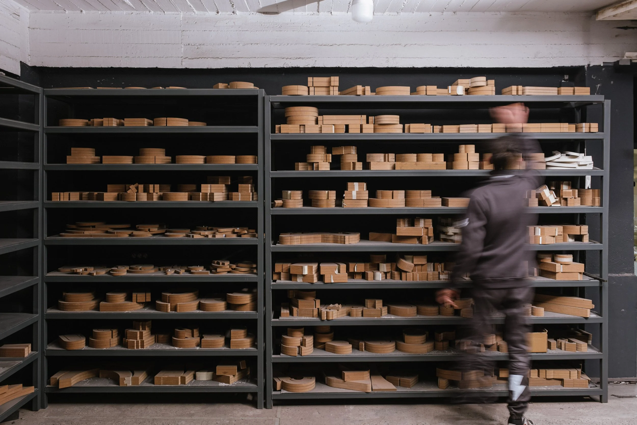 A person in motion walking past black metal shelves filled with various wooden pieces to be used in woodworking or manufacturing.