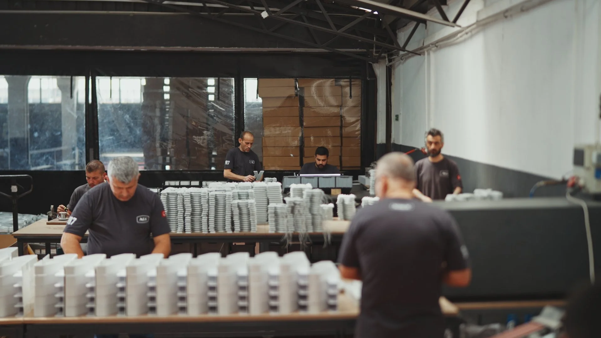 Workers in a factory assembling and inspecting white plastic trays or containers, with stacks of similar items on tables, in an industrial setting.
