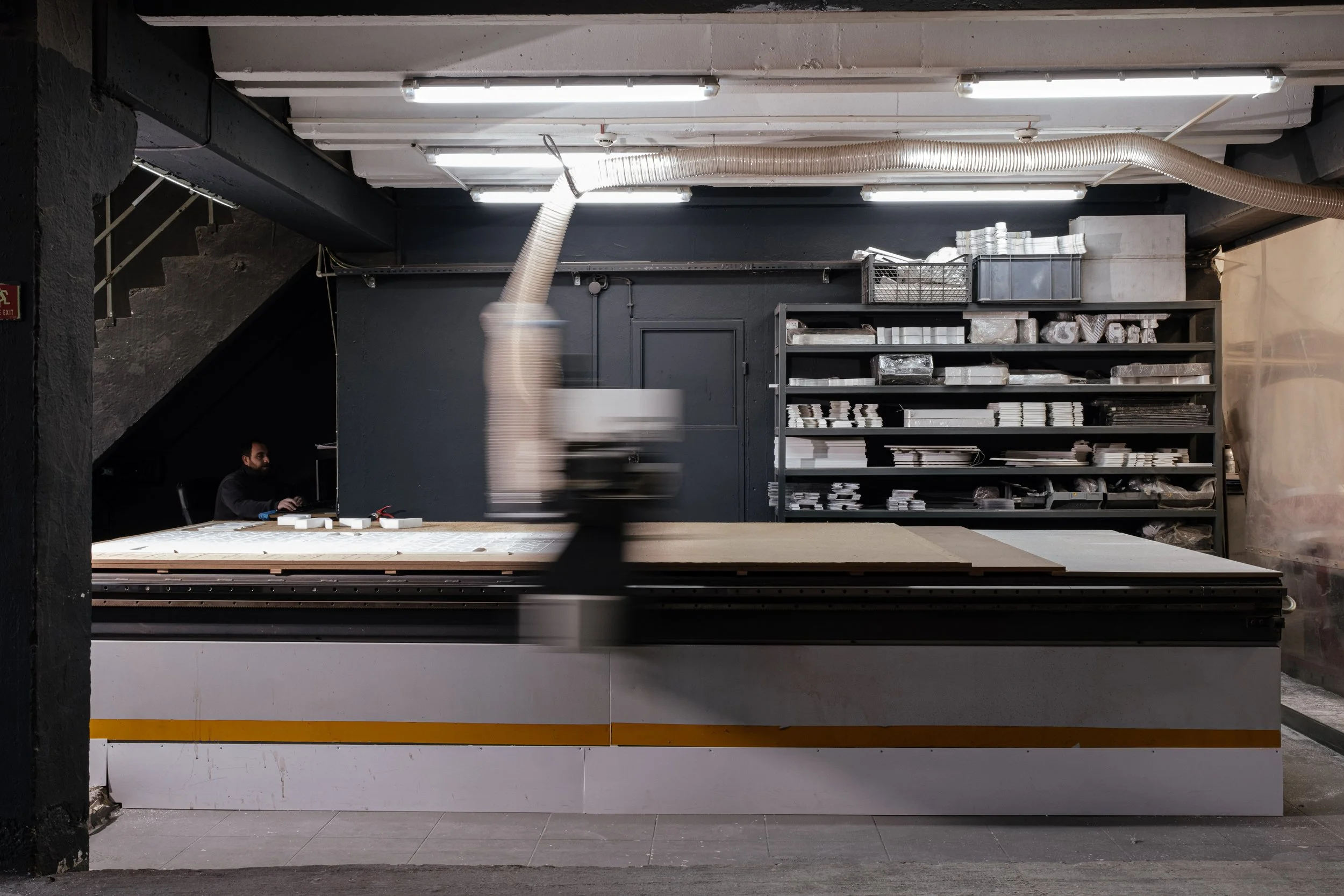 A workshop with a large cutting table, black shelving filled with materials, and a worker operating a machine with a pipe attached, in an industrial-style space with exposed ventilation and lighting.