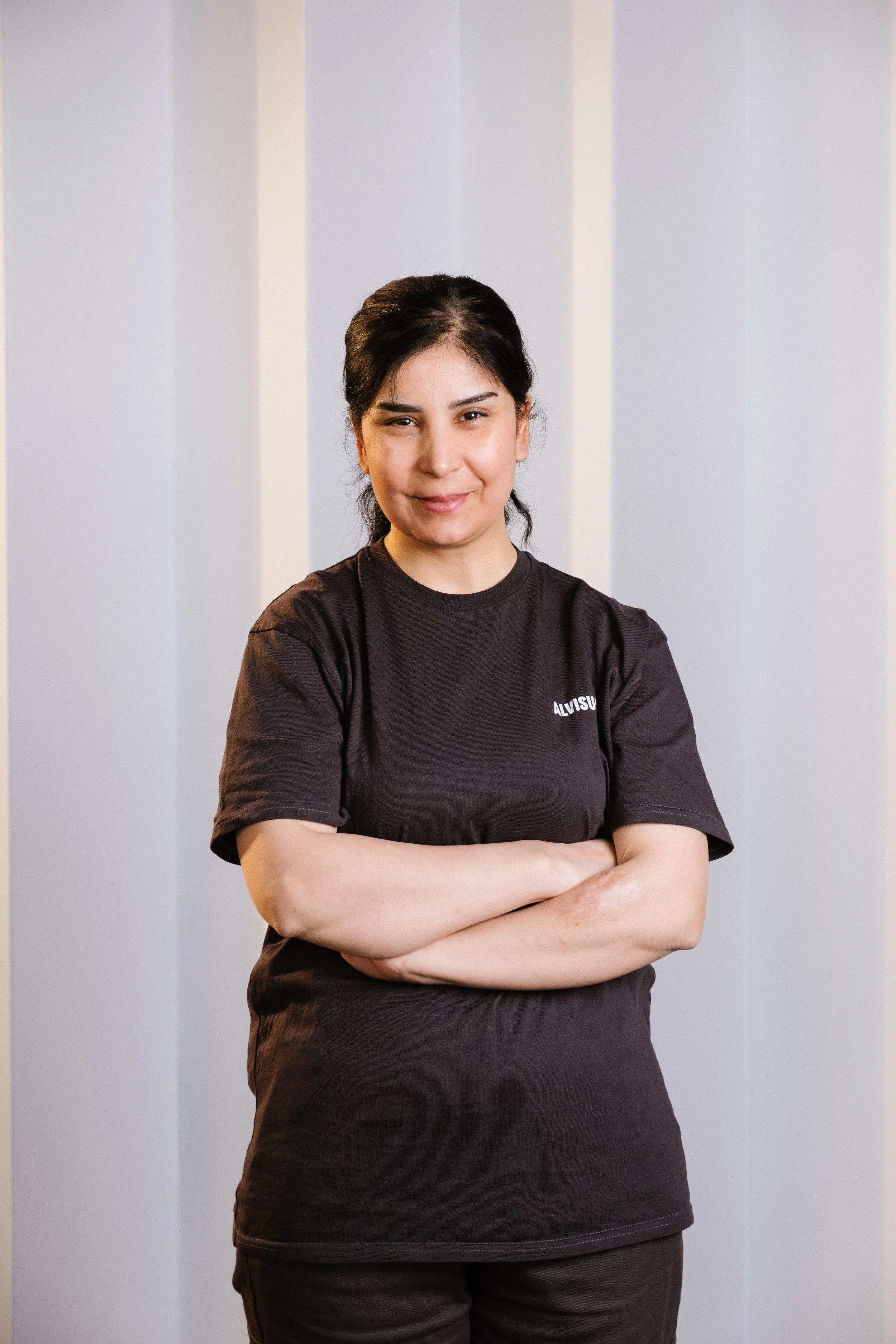 A woman with dark hair wearing a black T-shirt with white text, standing with her arms crossed in front of a light striped background.
