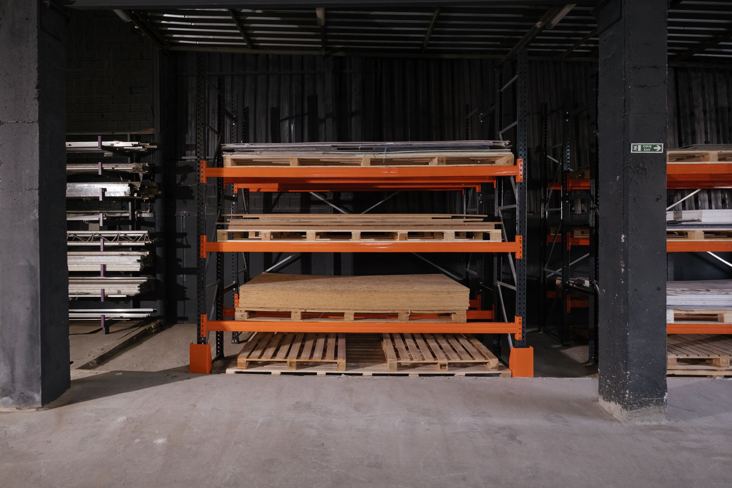 Industrial warehouse shelving with wooden pallets and construction materials stored on orange and black metal racks.