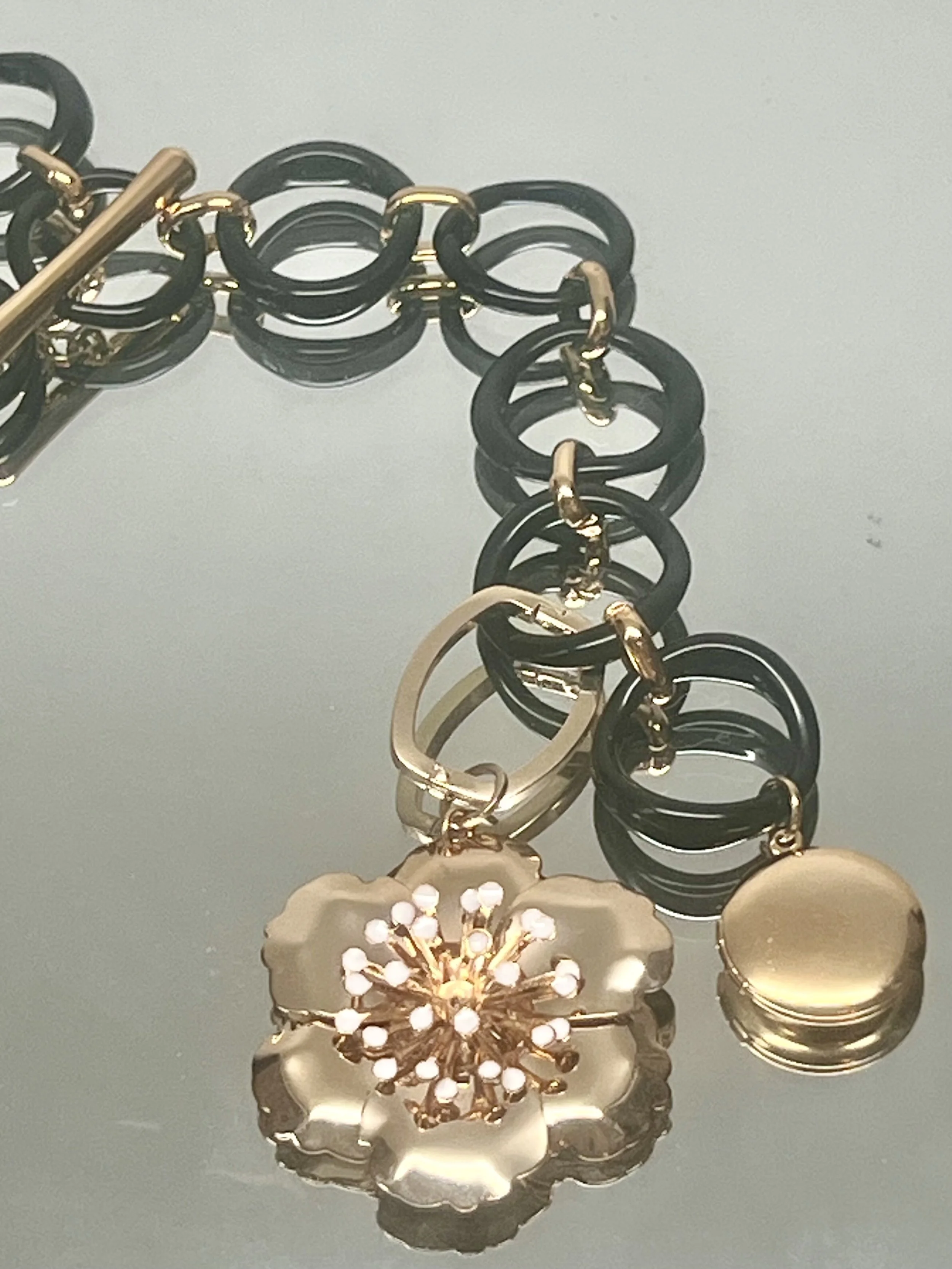 Close-up of jewelry, including a flower-shaped pendant with white stones, and black and gold rings and links, reflected on a smooth surface.
