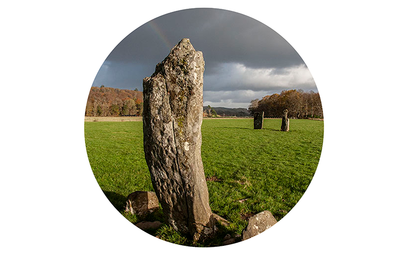 Standing ancient stone monument in a field with cloudy sky and a faint rainbow, with other stone markers in the distance.